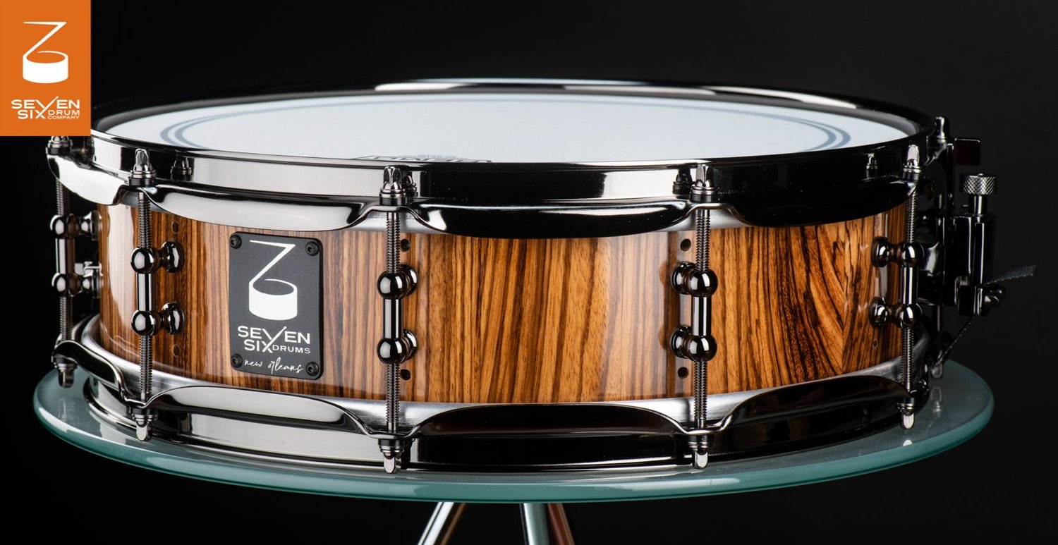 Handmade snare drum featuring a glossy zebrawood shell, black nickel hoops, and double-ended lugs.