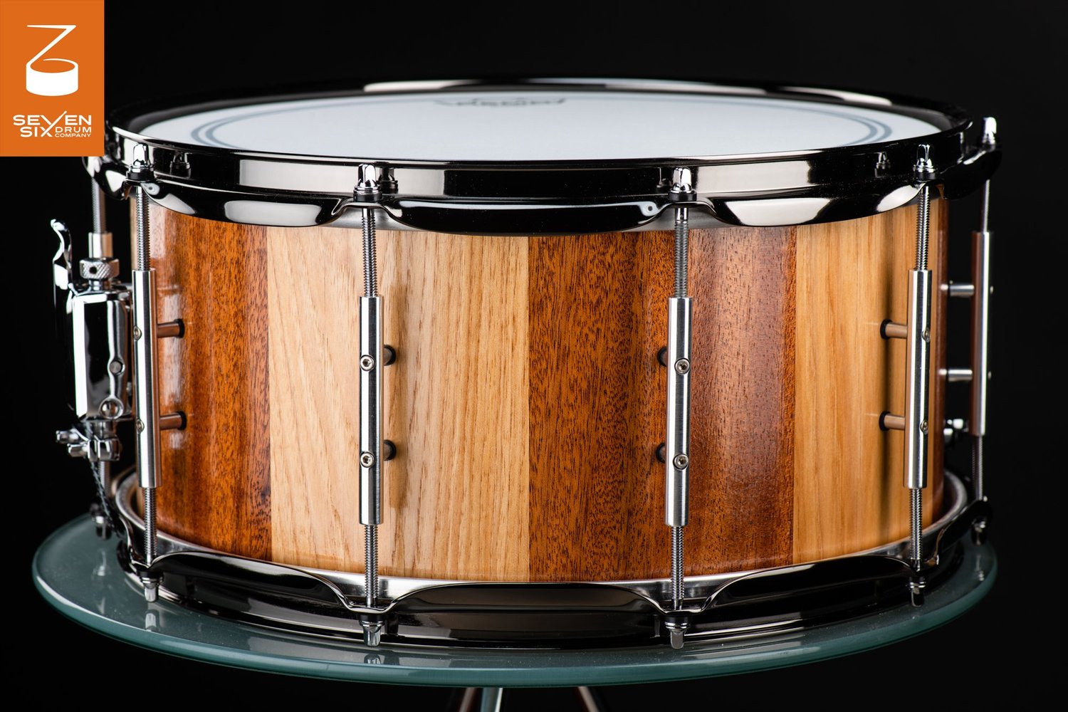 Handmade snare drum with alternating light and dark wood staves in a clear gloss finish, chrome tube lugs, and dark metal hoops.
