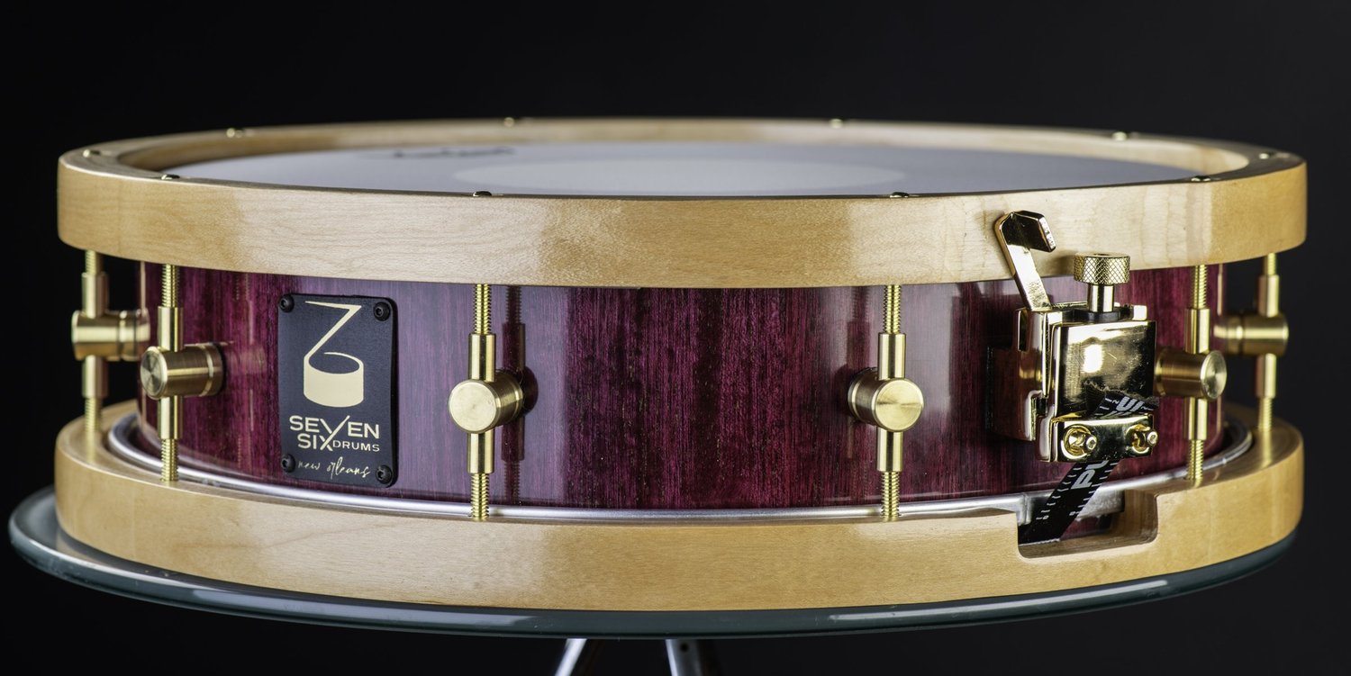Handmade snare drum with a glossy purple wood shell, natural blonde wood hoops, brass lugs, and a polished gold throw-off.