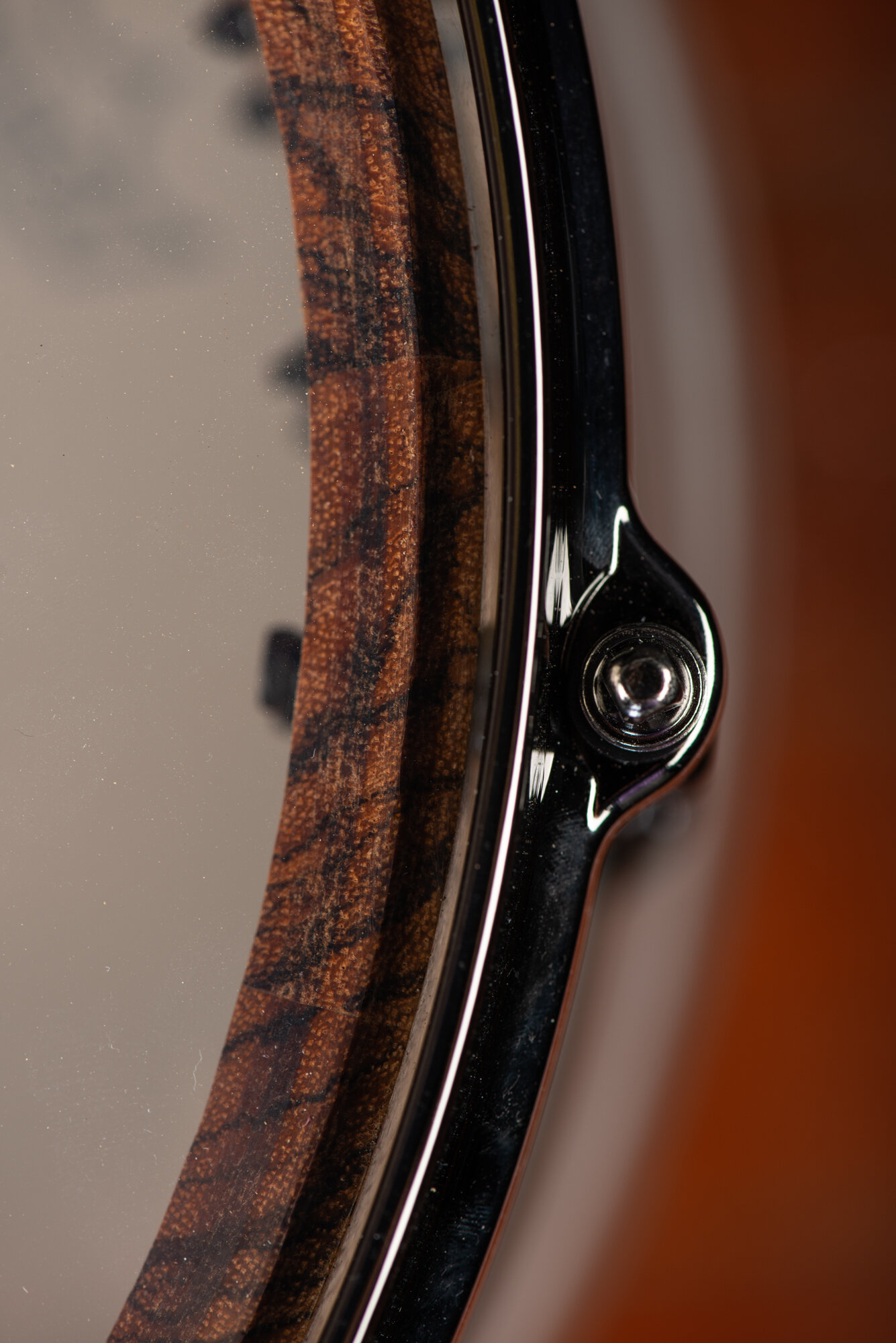 Close-up of a snare drum with a polished chrome hoop, tension bolt, and figured dark wood shell visible through the drumhead.