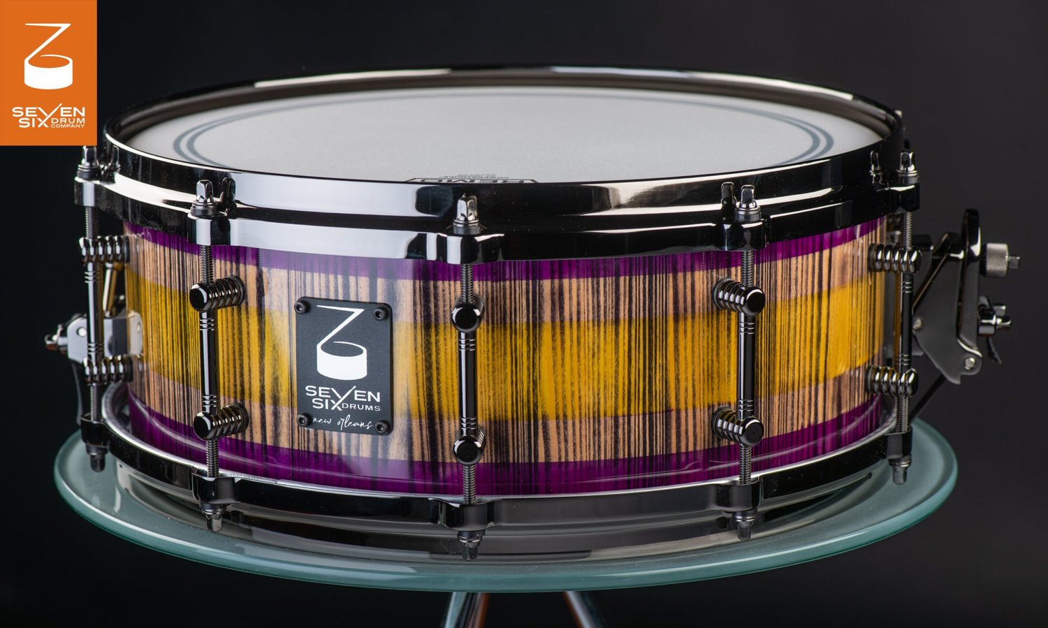Custom Louisiana Cypress snare drum featuring a purple and yellow striped wood grain finish with black nickel tube lugs and hoops.