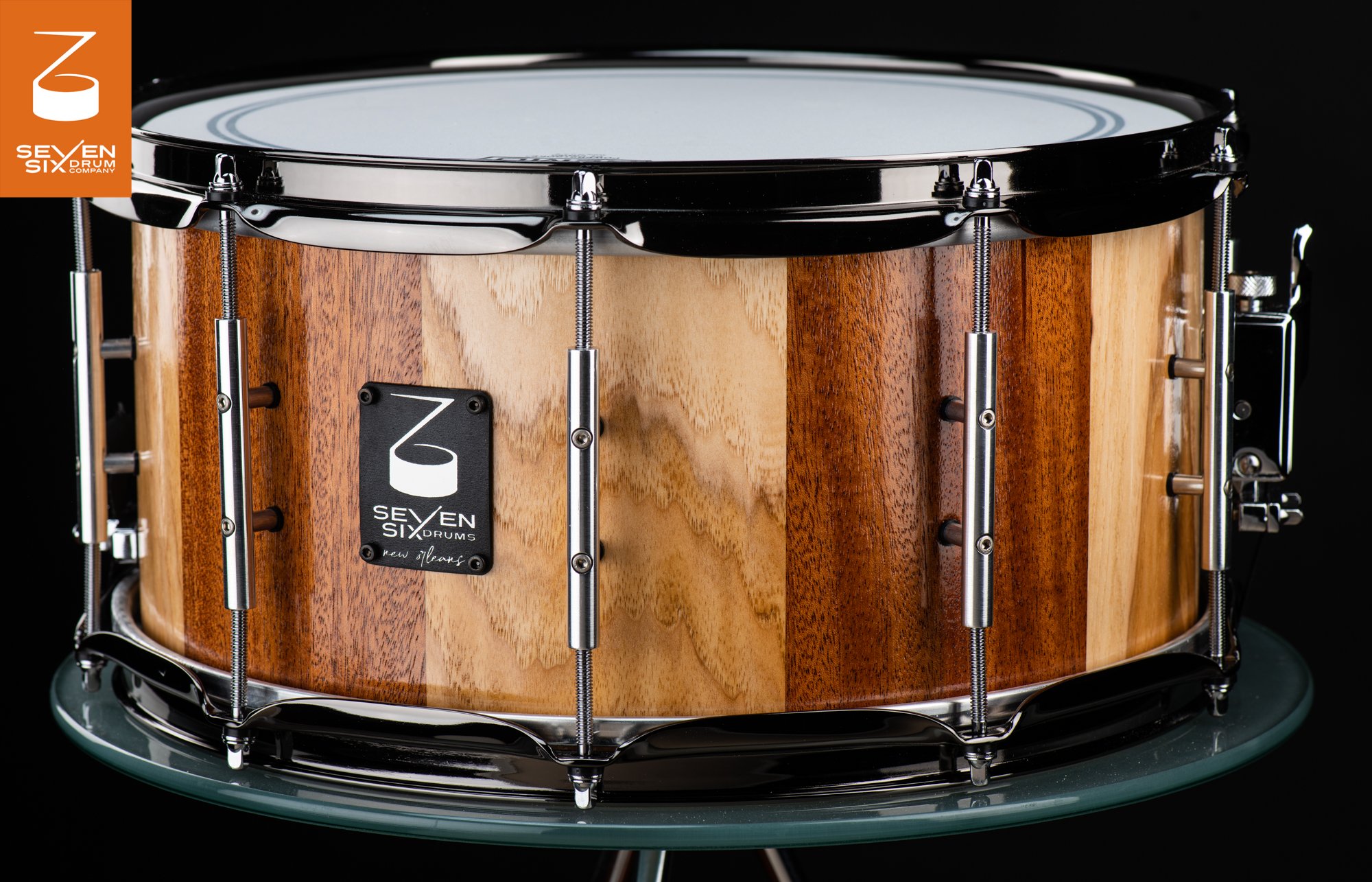 Handmade snare drum with alternating light and dark wood staves, a gloss finish, chrome tube lugs, and metal hoops.