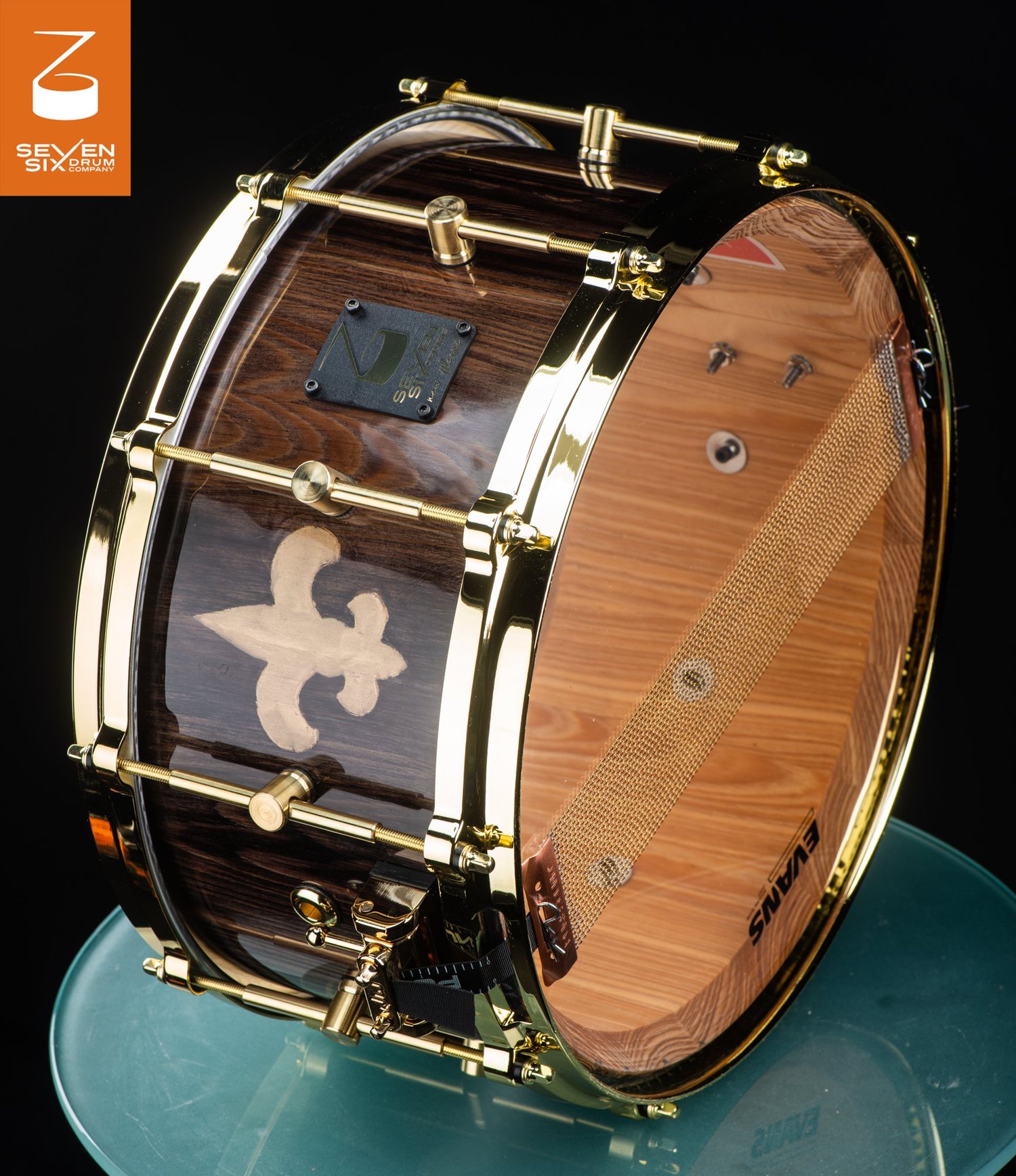 Handmade snare drum with a dark gloss wood shell, fleur-de-lis inlay, and polished gold hoops and tube lugs.