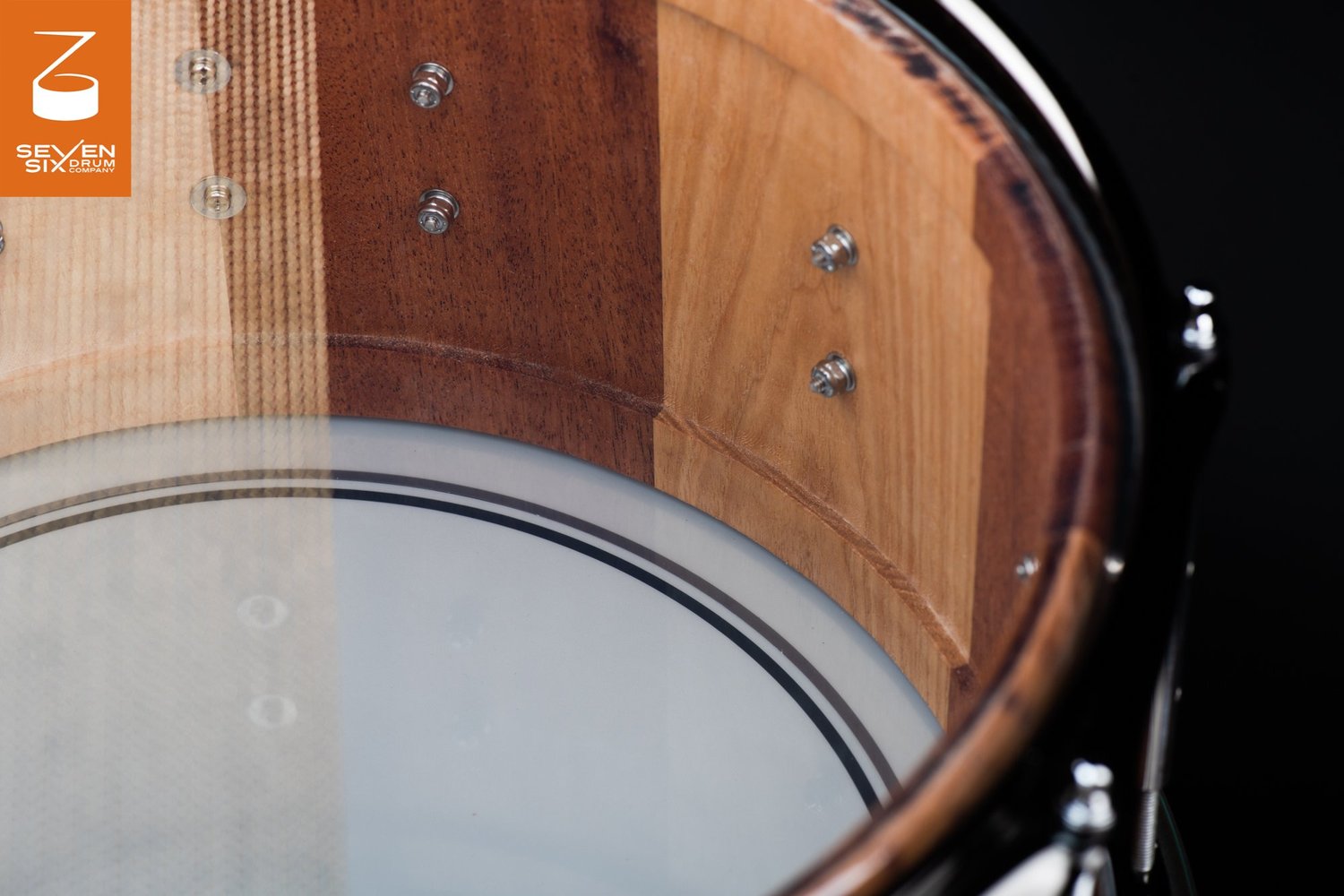 Interior of a stave snare drum shell with alternating light and dark natural wood staves and chrome lug mounting bolts.
