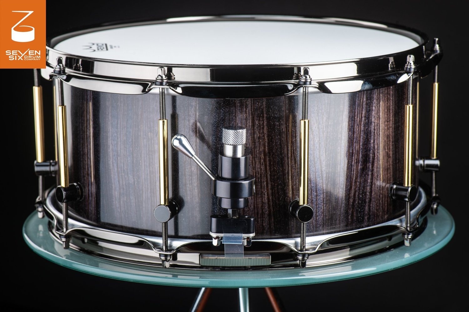 Handmade snare drum featuring a glossy dark wood shell, black nickel hoops, and brass-accented tube lugs.