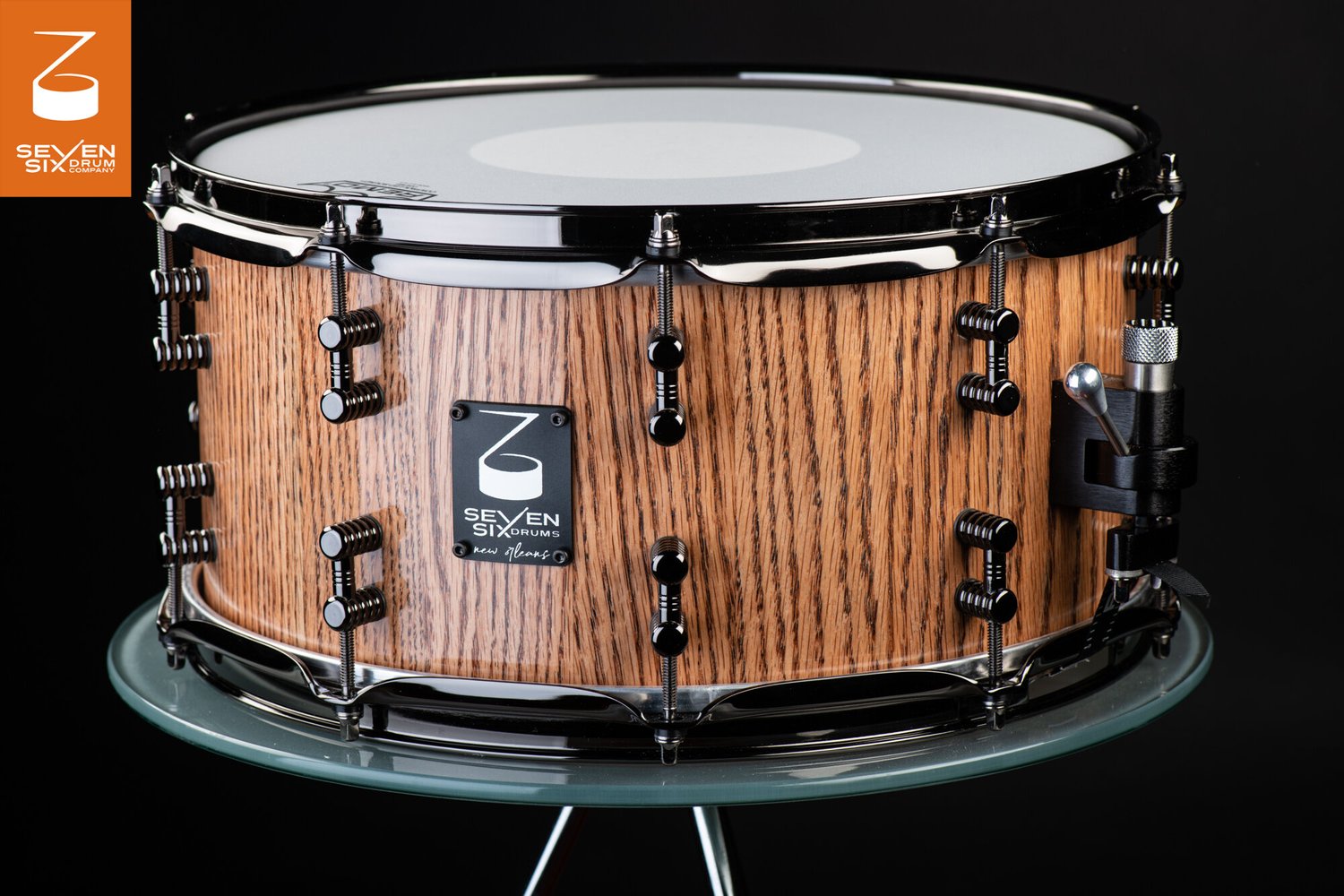 Handmade snare drum with a natural oak wood finish, black nickel hoops, segmented black lugs, and a throw-off.