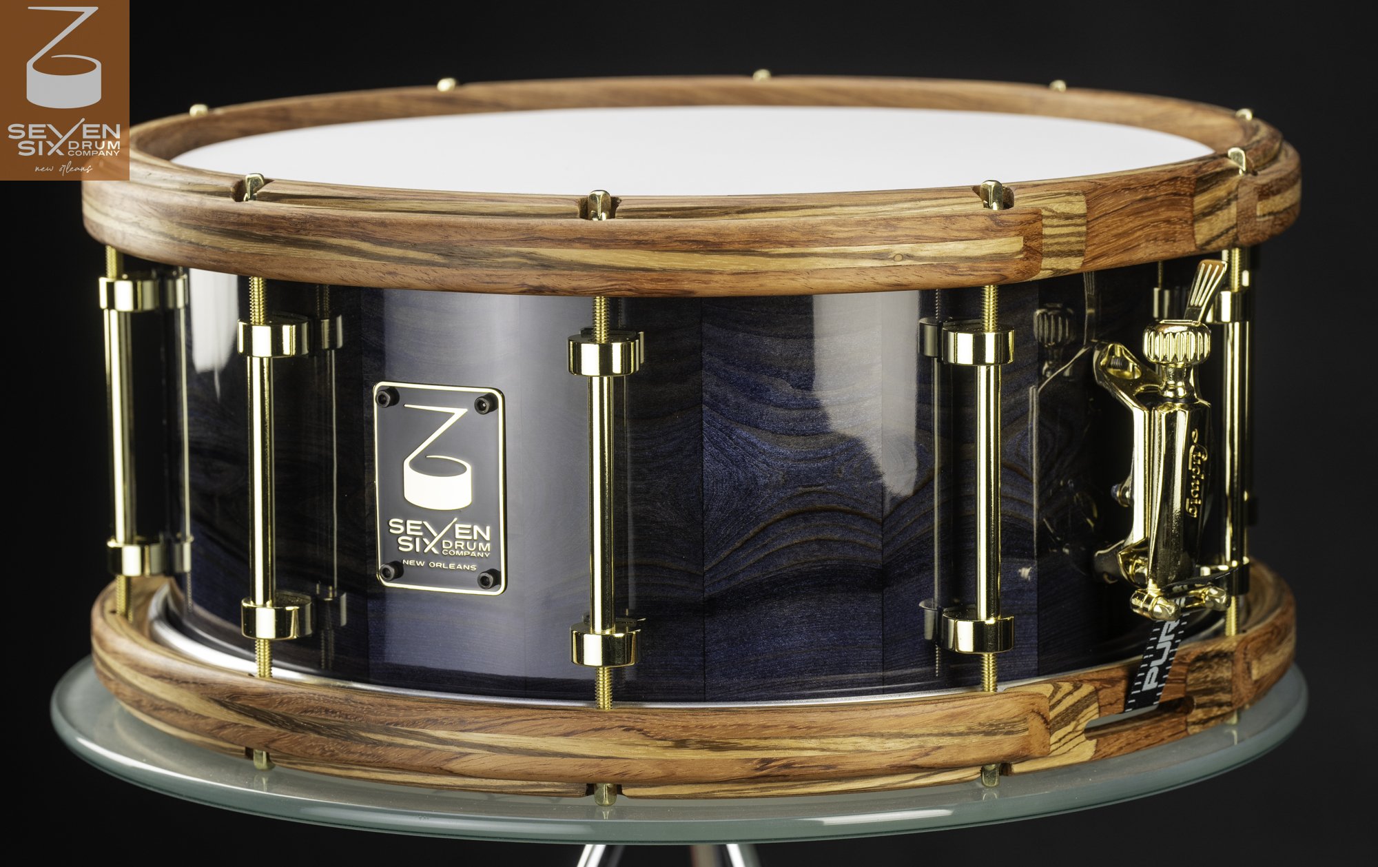Handmade snare drum with a glossy blue wood shell, striped wooden hoops, and polished gold tube lugs and throw-off.