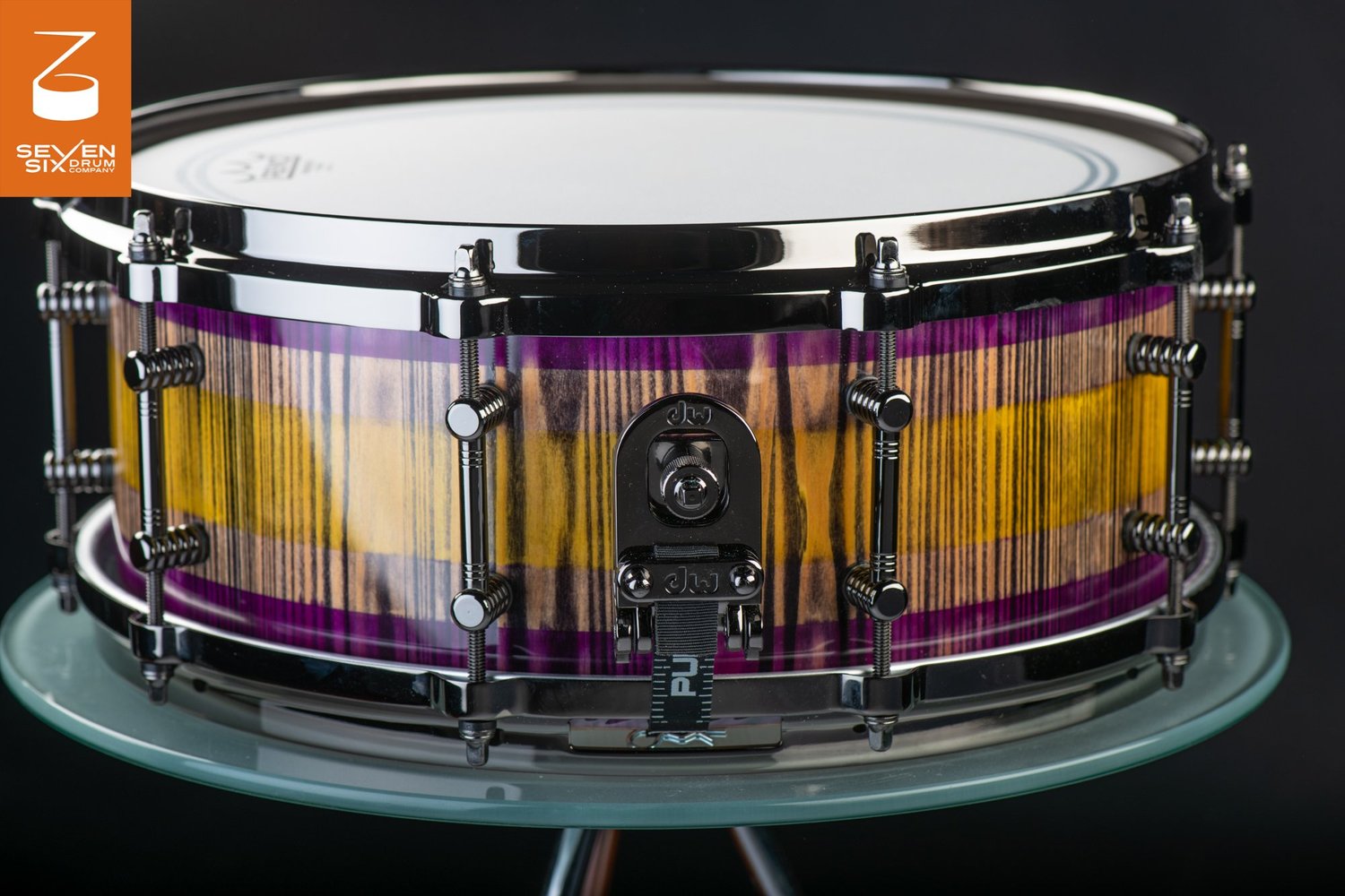Handmade snare drum with a purple and yellow striped wood finish, black nickel hardware, and a DW butt plate.