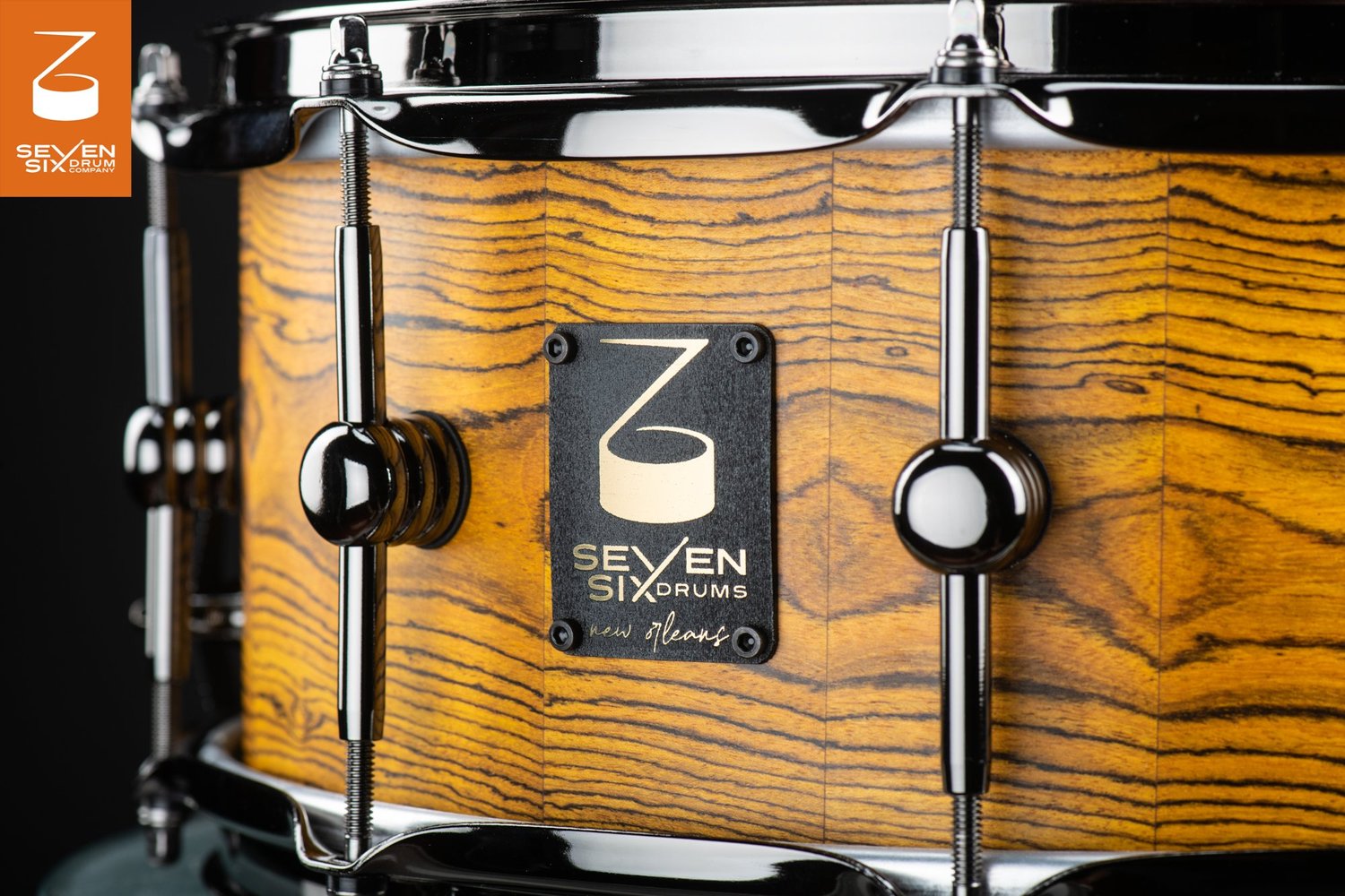 Snare drum with a golden natural wood shell, black nickel lugs and hoop, and a black Seven Six Drums badge.