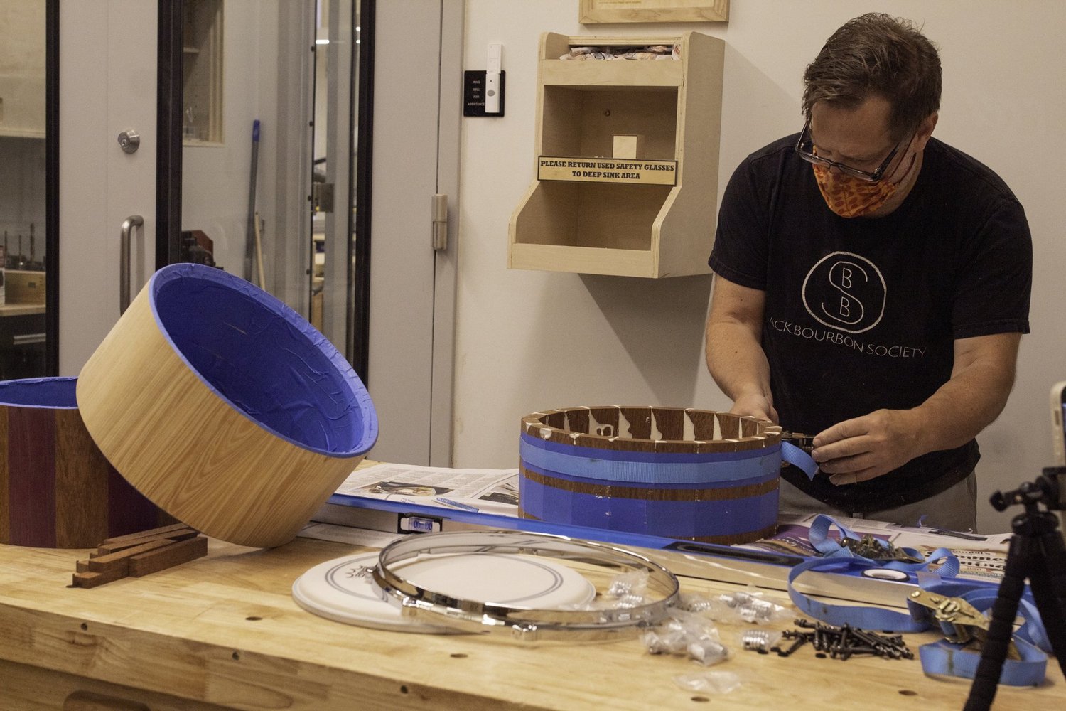 A drum maker clamps a segmented wooden shell on a workbench beside an unfinished stave shell and chrome hoops.