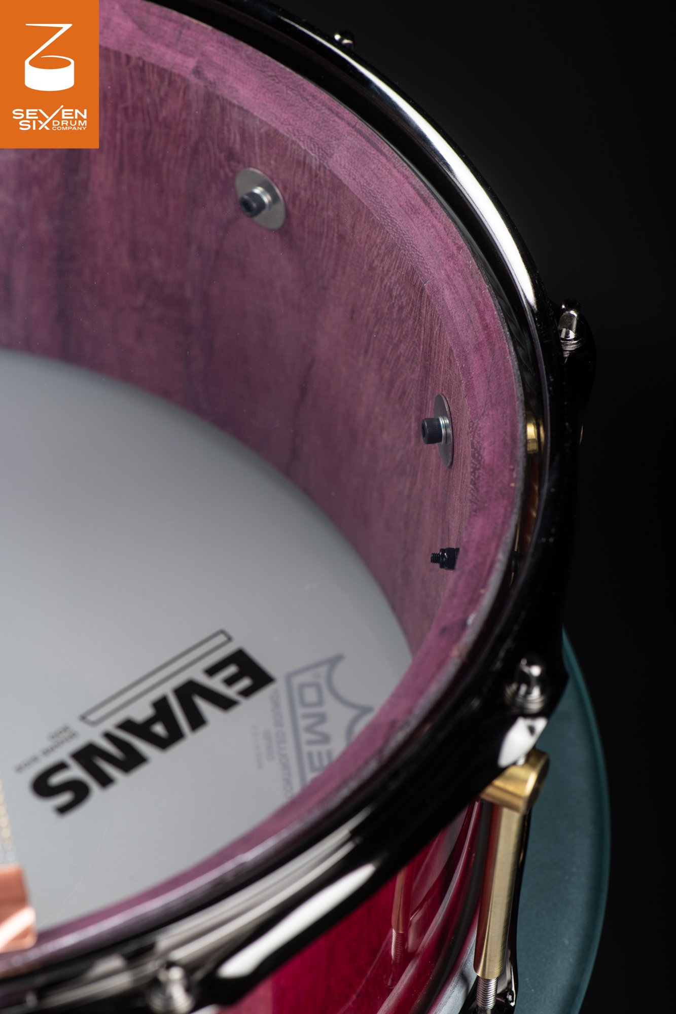 Interior of a purpleheart wood snare drum showing the natural purple grain, black drum hoop, and brass tube lugs.