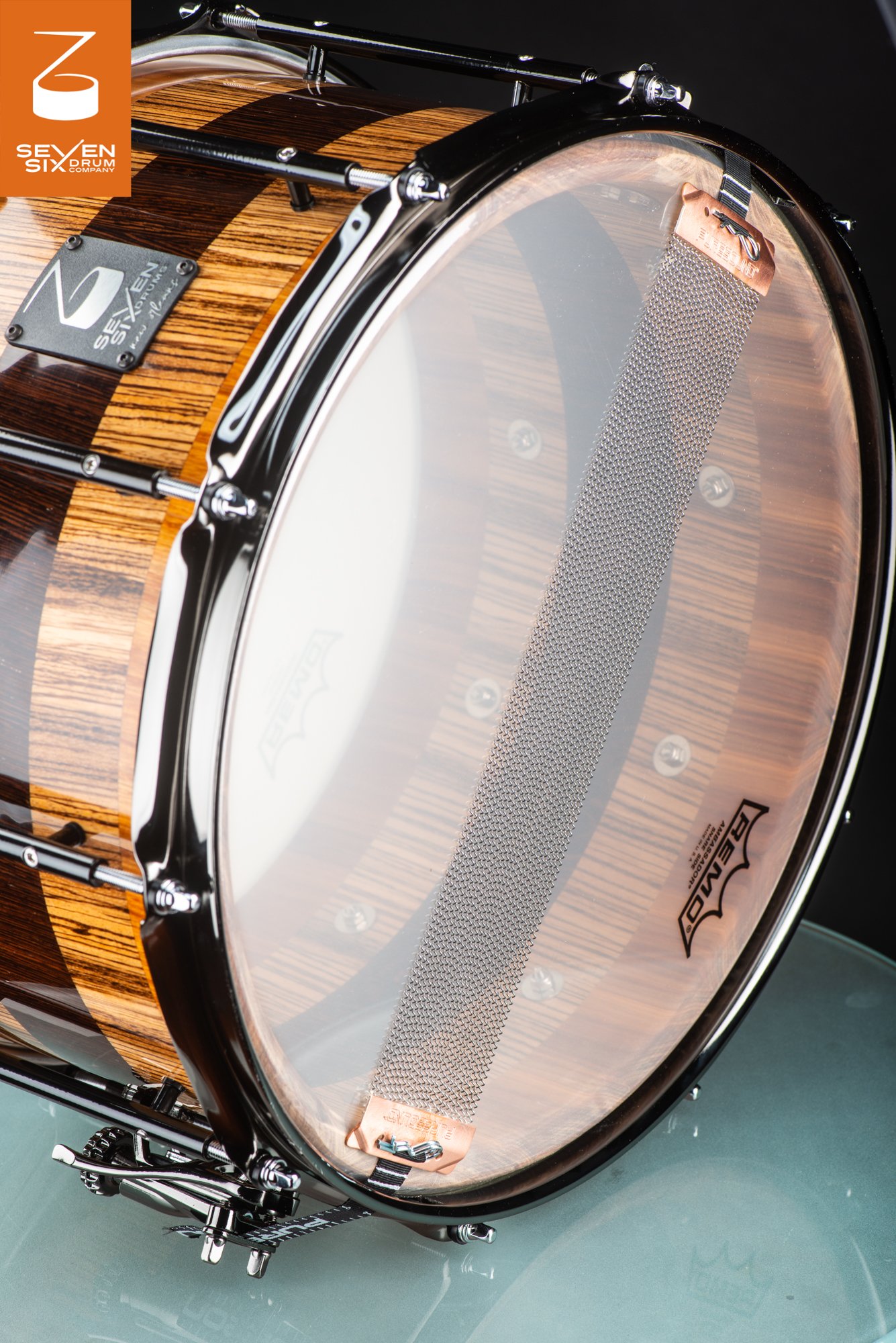 Underside of a glossy striped wood snare drum with a clear resonant head, wide snare wires, and black metal hardware.