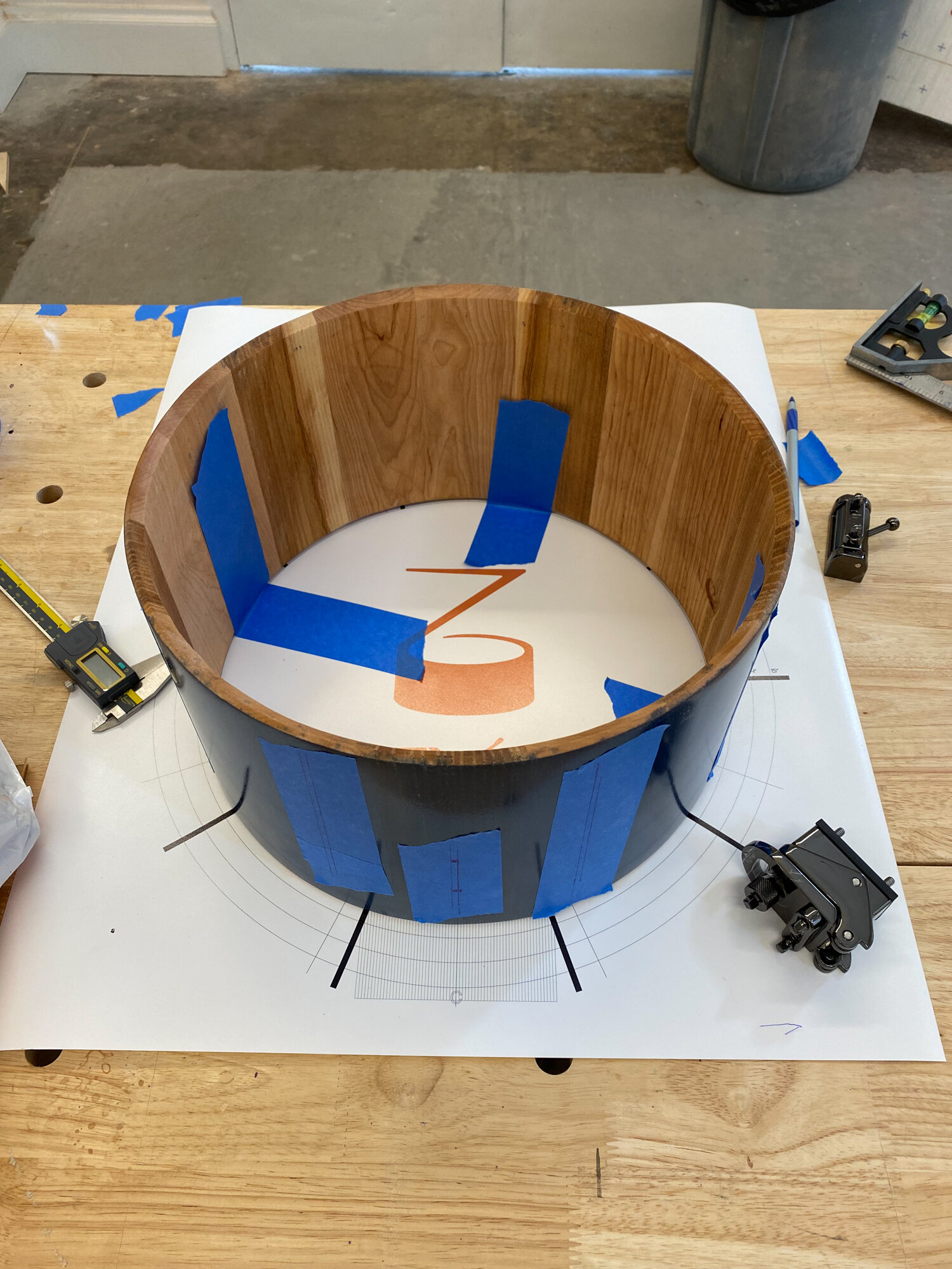 A dark stave wood snare drum shell with a natural interior sits on a layout mat beside an unmounted black snare strainer.
