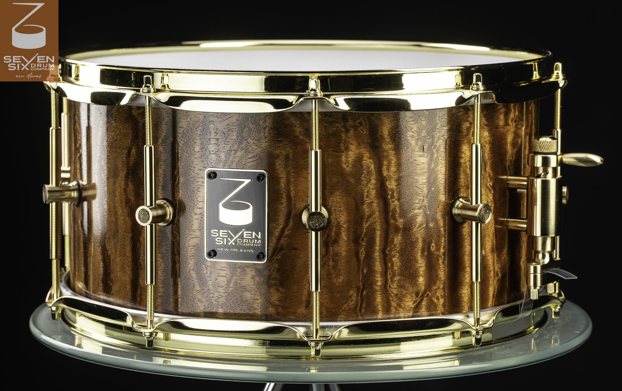 Handmade snare drum with a glossy figured brown wood shell, polished brass hoops, and single-point brass lugs.
