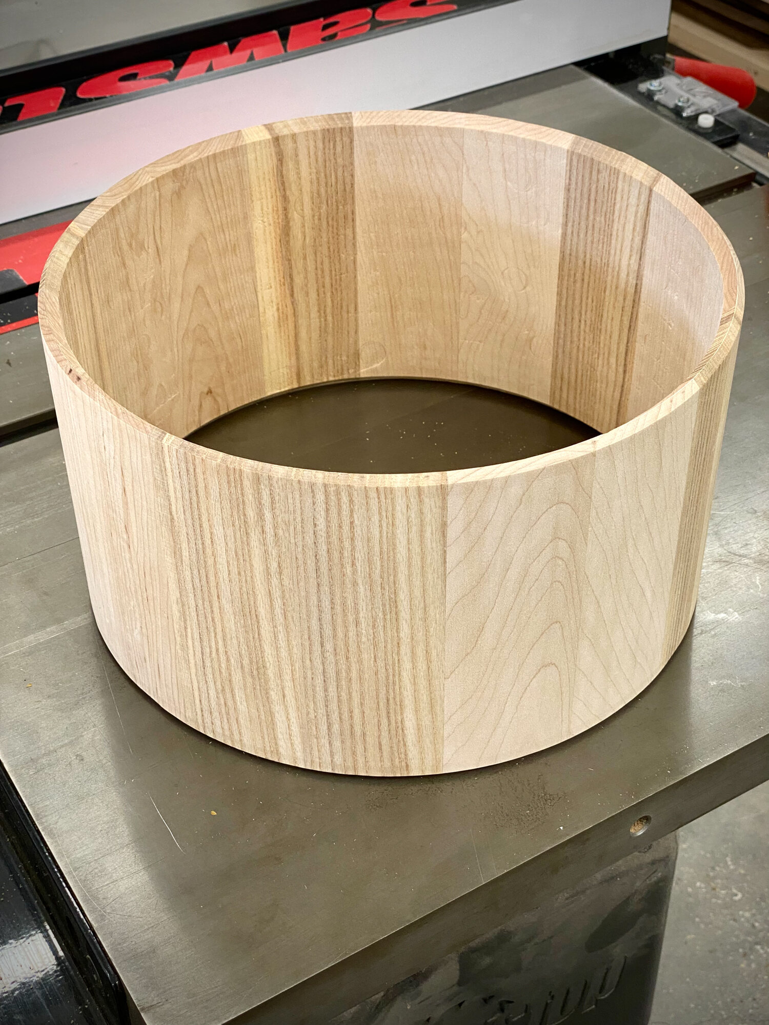 An unfinished stave snare drum shell made of raw light wood with smooth bearing edges and no hardware installed.