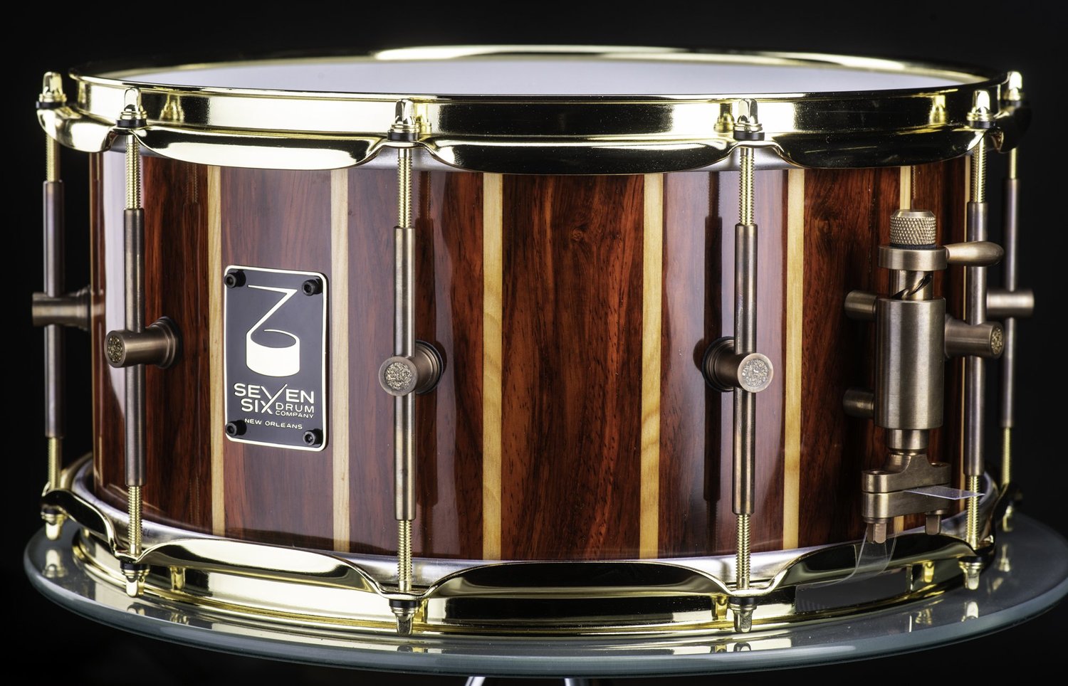 Gloss-finish striped Padauk and Louisiana Cypress wood snare drum featuring red and blonde vertical staves, polished brass hoops, and bronze lugs.