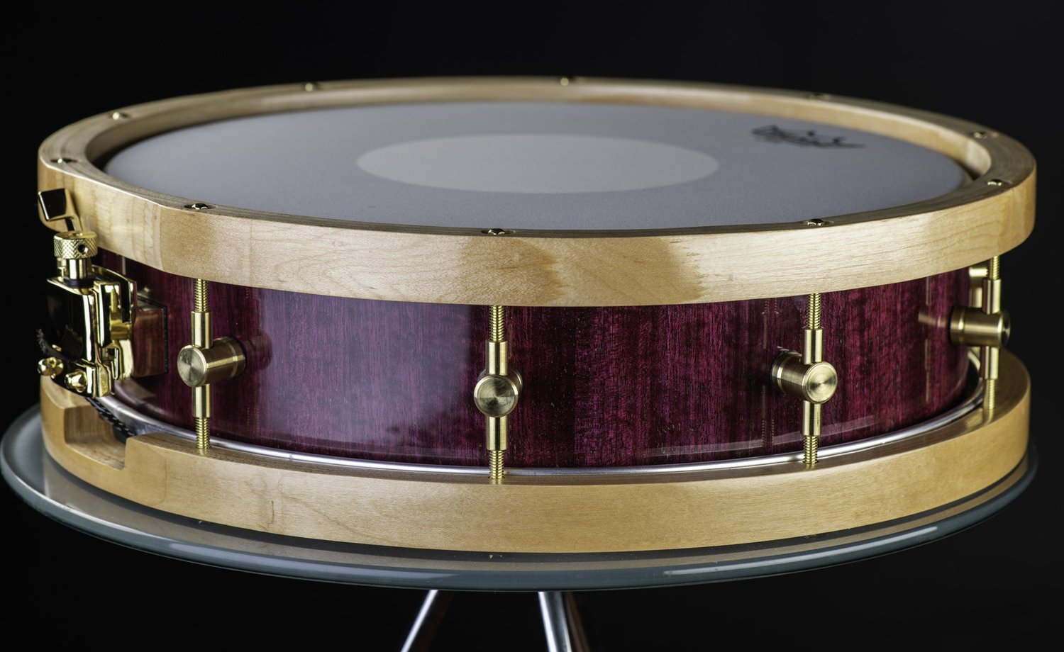Handmade snare drum featuring a glossy purple wood shell, natural blonde wood hoops, and polished brass hardware.
