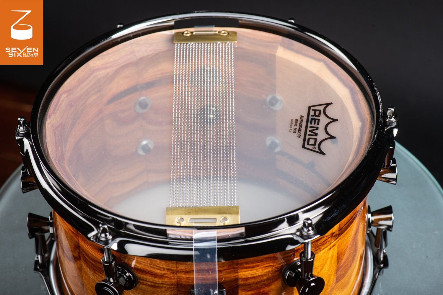 Snare-side view of a glossy natural wood snare drum fitted with black hoops, black lugs, and wire snares across a clear head.