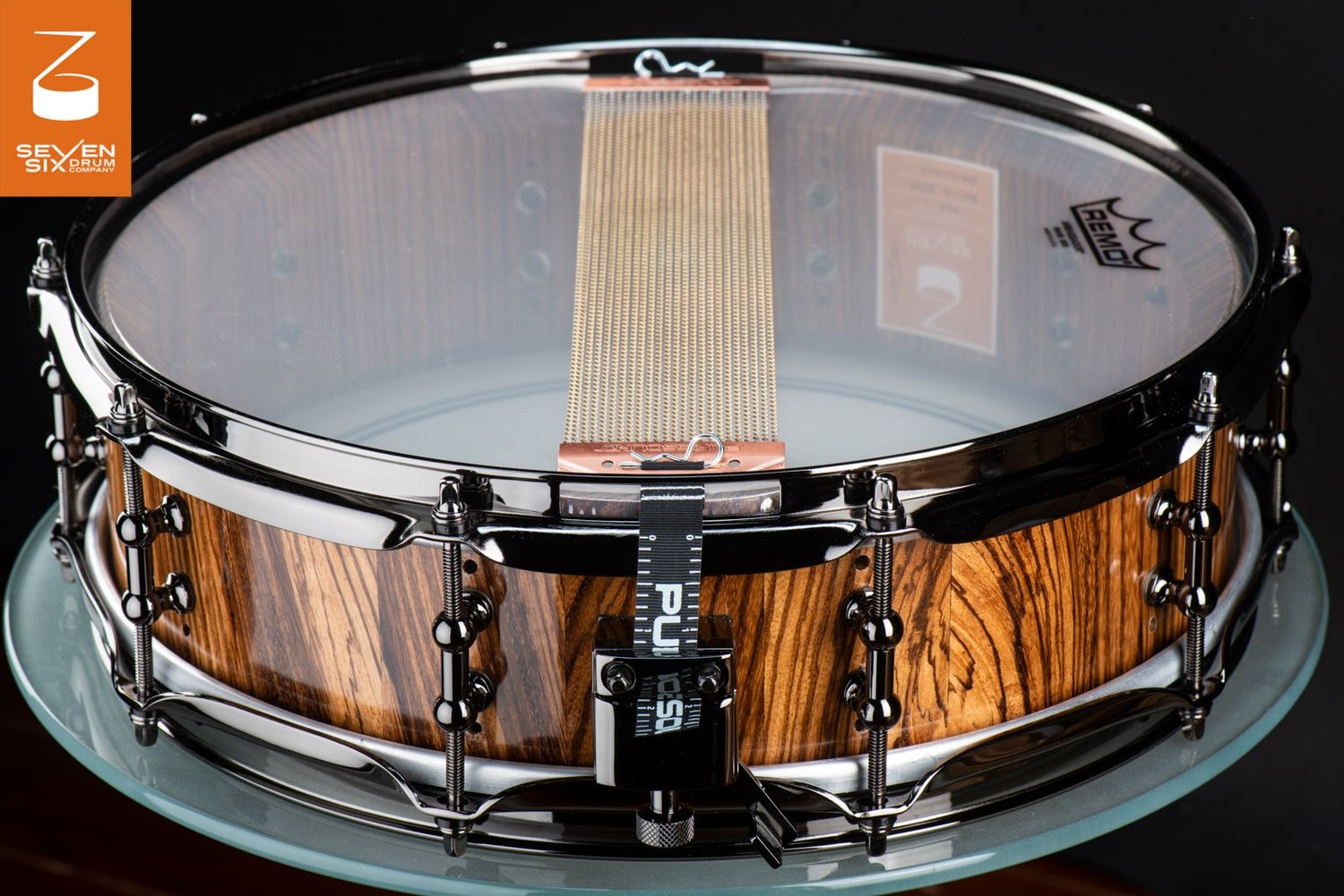 Handmade snare drum with a glossy zebrawood shell, black nickel lugs and hoops, and snare wires over a clear head.
