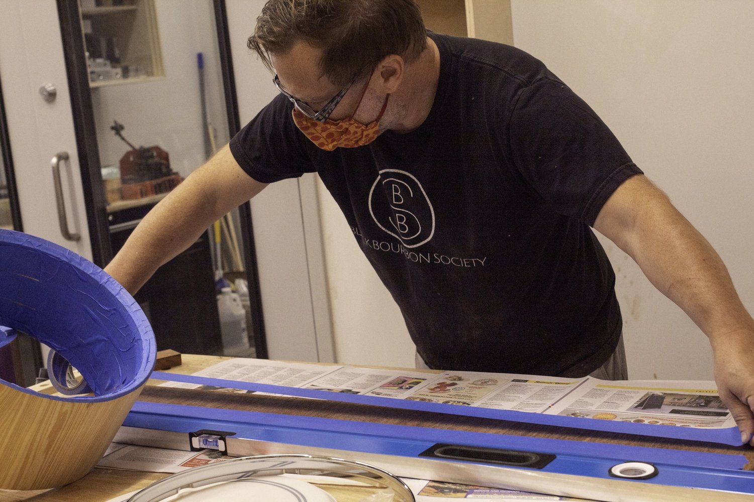 A drum maker tapes a dark wood veneer beside an unfinished natural wood shell and a polished chrome snare hoop.