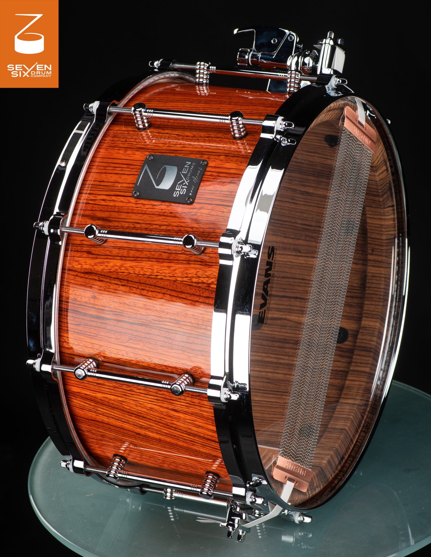 Handmade snare drum featuring a glossy reddish wood shell, chrome tube lugs, polished hoops, and visible bottom snare wires.