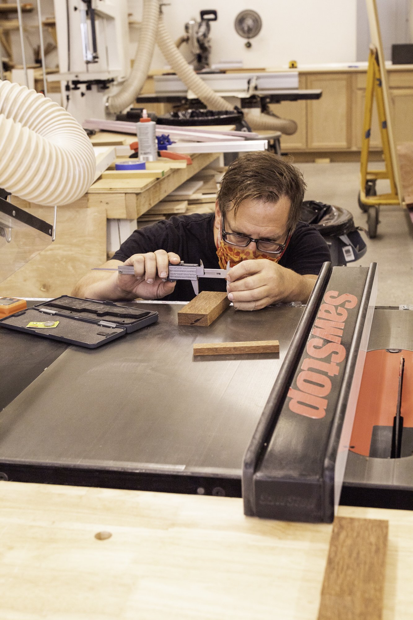 A drum maker measures a block of unfinished hardwood with metal calipers on a steel table saw.