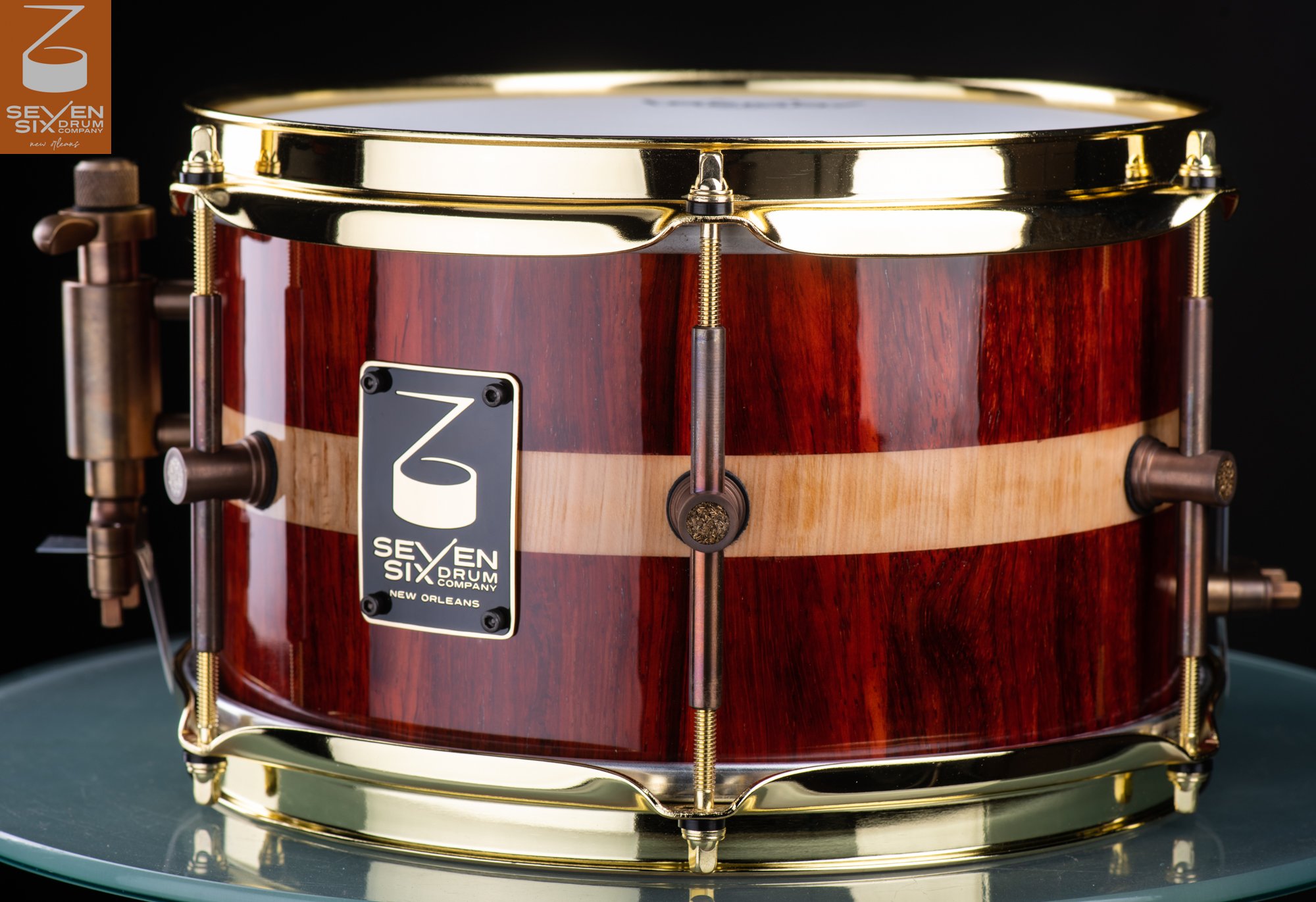 Handmade snare drum in glossy red-brown wood with a light center stripe, polished brass hoops, and antique bronze hardware.
