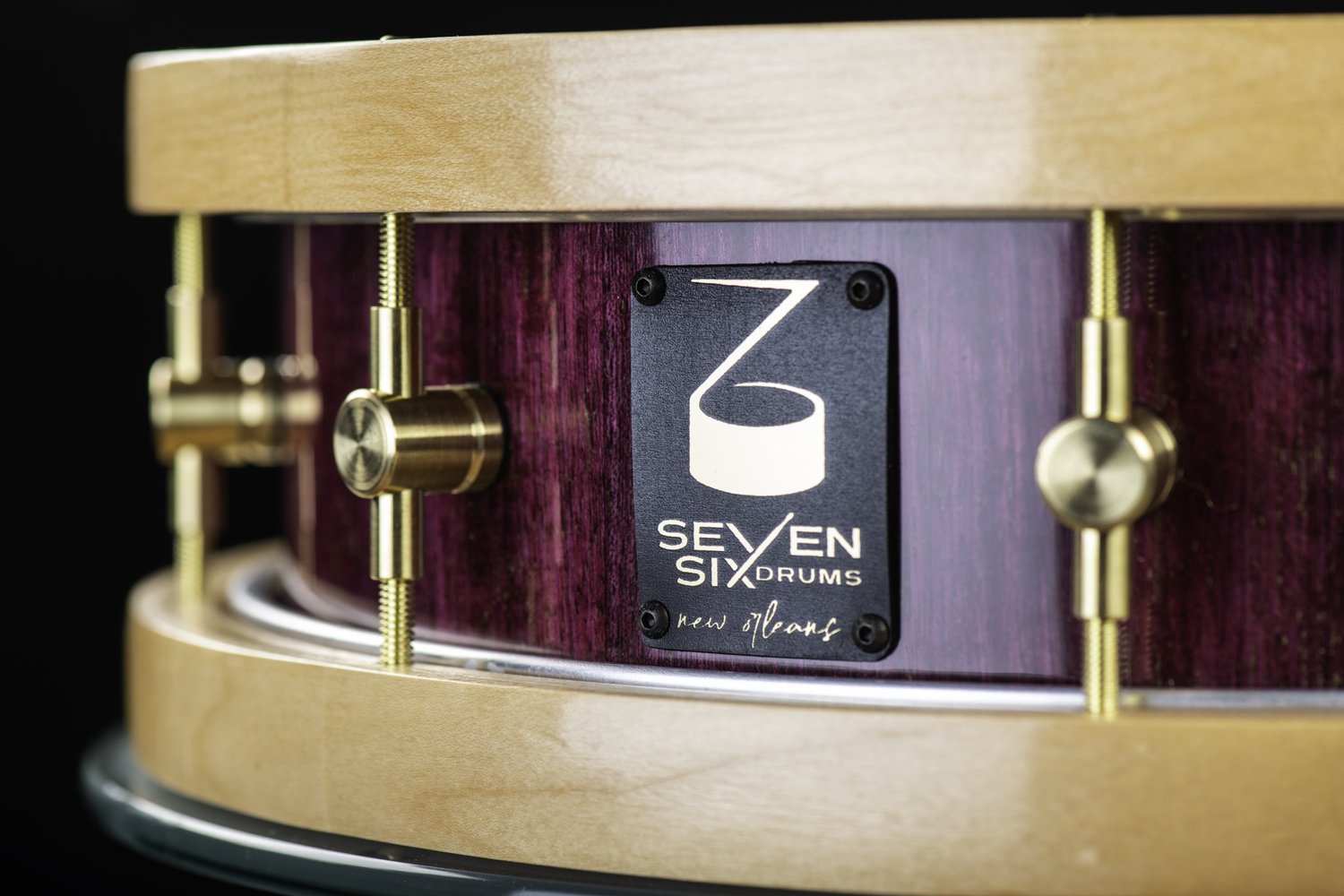 Glossy purple wood snare drum with natural wood hoops, brass tension lugs, and a black brand badge.