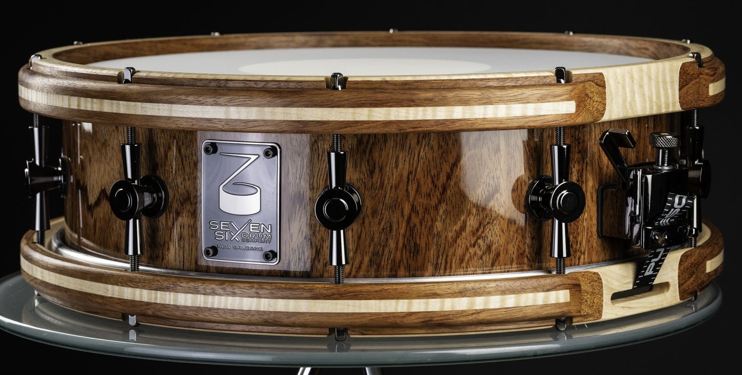 Handmade bubinga snare drum with a gloss wood shell, Bubinga with curly maple inlay wooden hoops, and black nickel hardware.