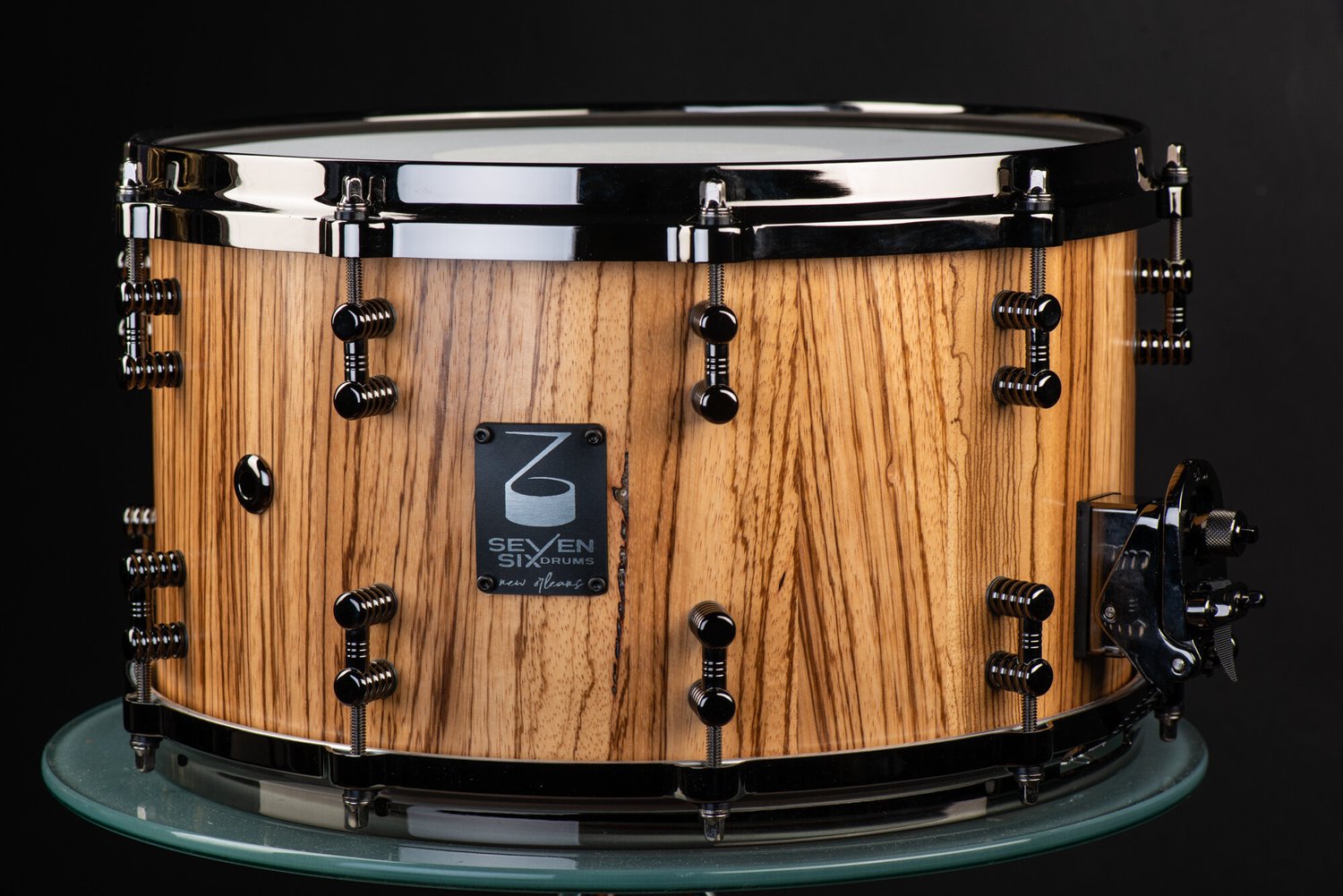 Handmade snare drum featuring a natural striped wood shell, black nickel hoops, and matching black segmented lugs.