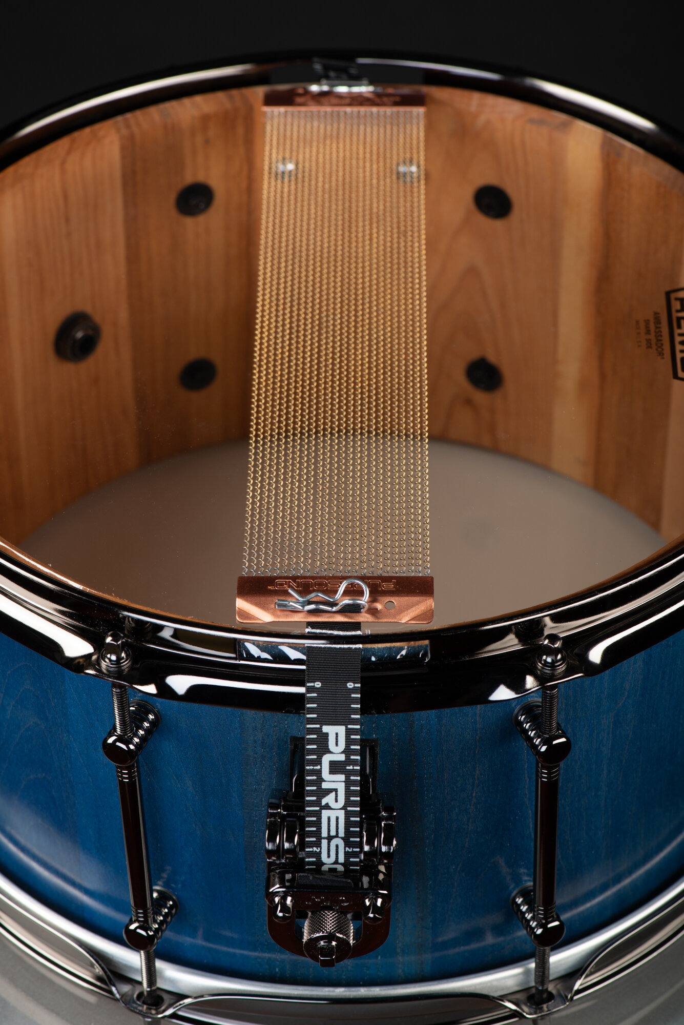 Blue-stained wooden snare drum with black hardware, brass snare wires, and a clear head showing the natural interior.