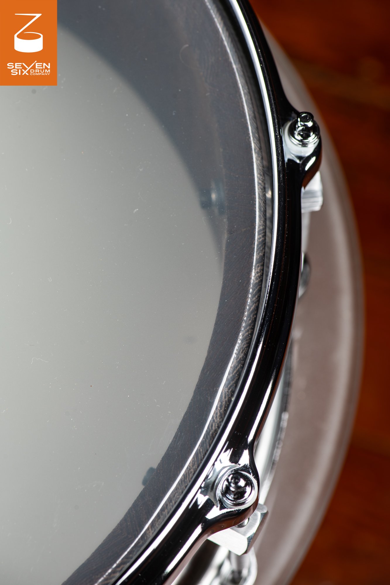 Detail of a snare drum featuring a dark wood shell, clear batter head, and a polished chrome rim with tension rods.