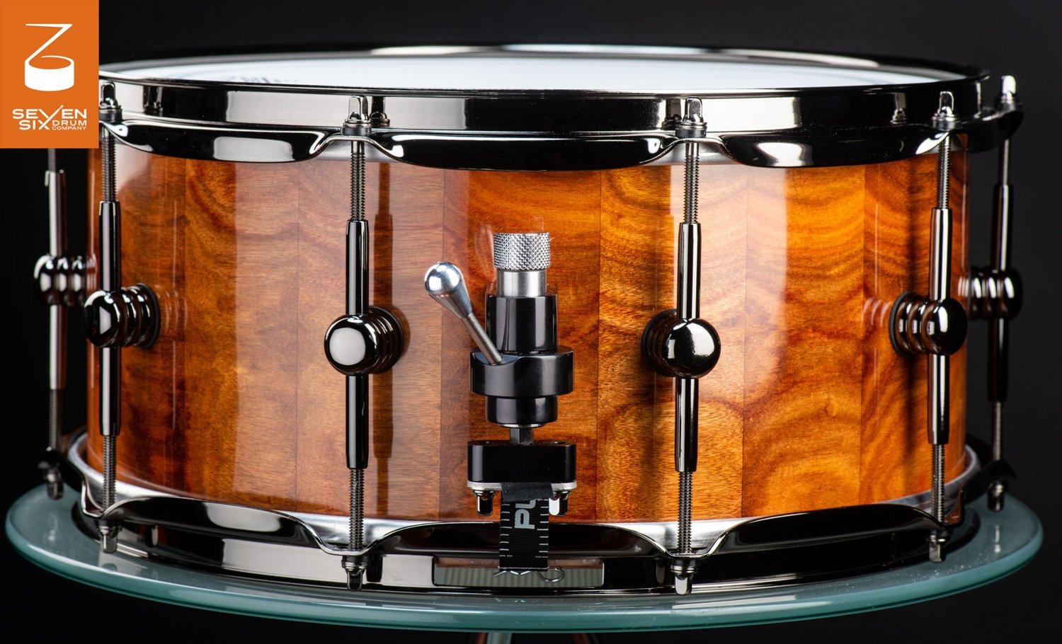 Handmade snare drum with a glossy figured wood shell, black nickel hoops and lugs, and a snare throw-off.