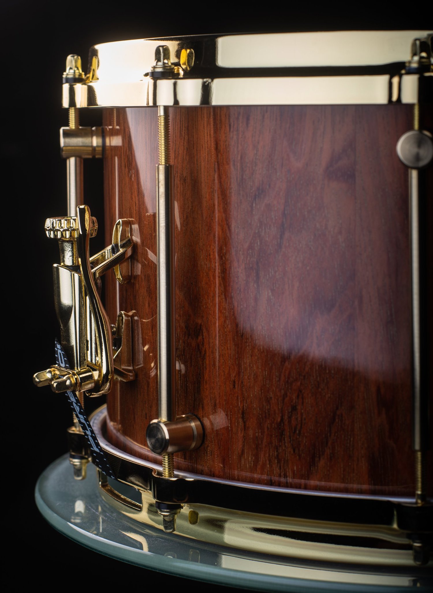 Handmade snare drum with a high-gloss brown wood shell, polished gold hoops, tube lugs, and a snare throw-off.