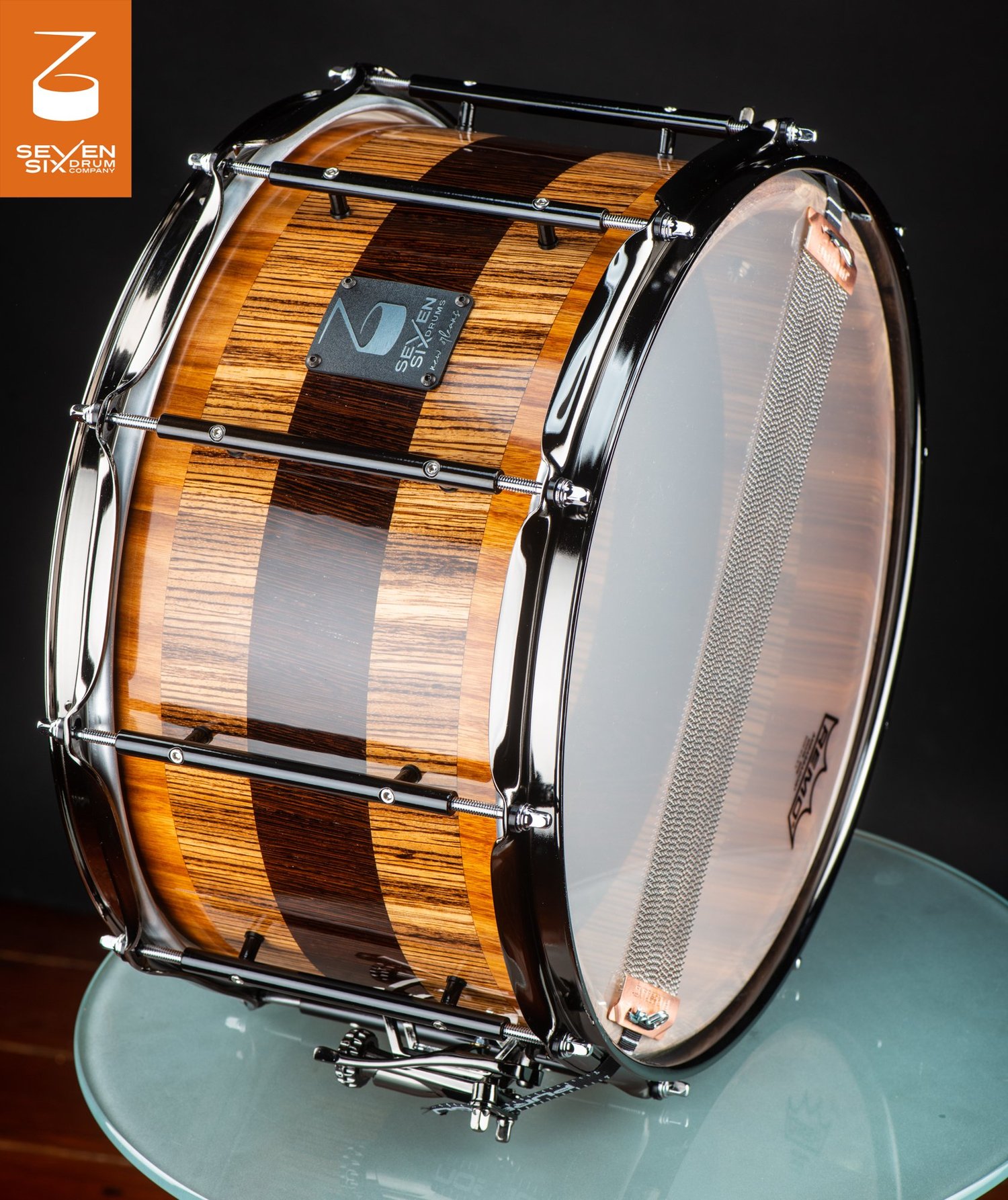 Glossy striped wood snare drum featuring black tube lugs, chrome hoops, and snare wires visible through a clear bottom head.