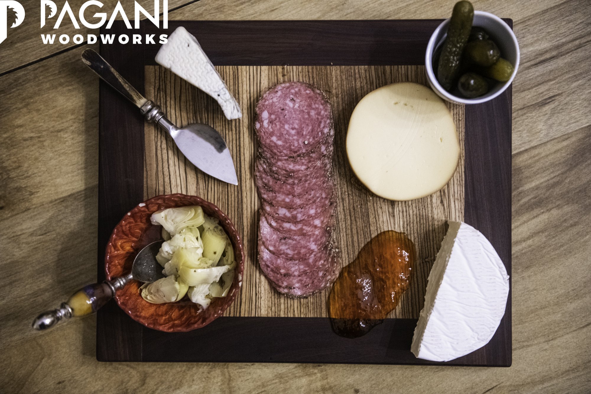Two-tone walnut and oak serving board with sliced salami, cheeses, preserves, and a vintage metal cheese knife.