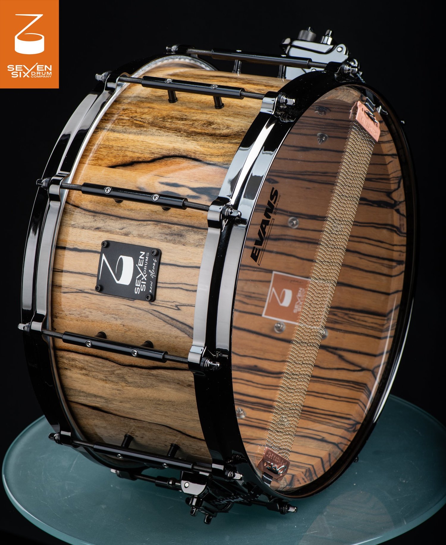 Handmade snare drum in glossy striped natural wood, featuring black hoops, tube lugs, and brass snare wires.