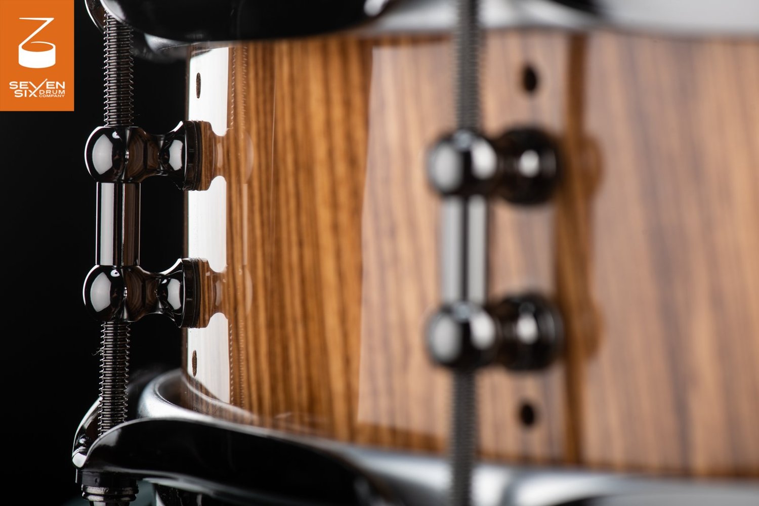 Close-up of a snare drum with a high-gloss wood grain shell, black nickel tube lugs, and tension rods.
