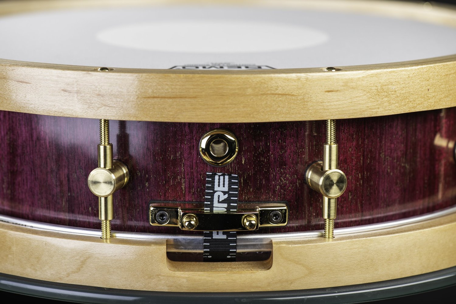 Purple-stained wood snare drum featuring natural blonde wood hoops, brass tension lugs, and a gold butt plate.