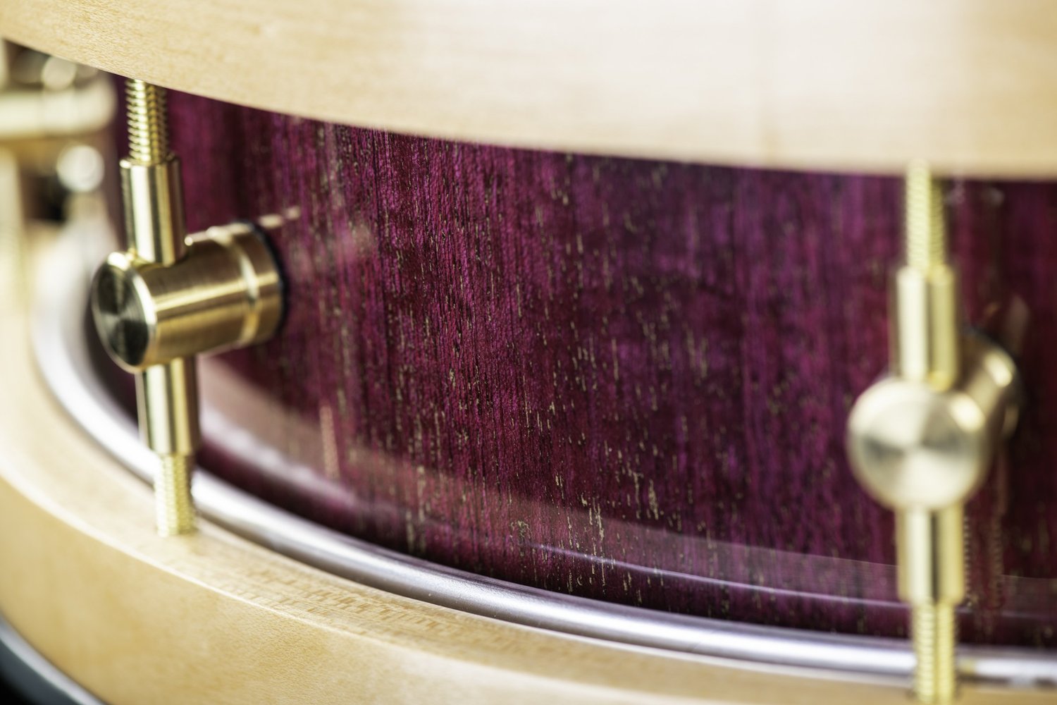 Close-up of a snare drum featuring a gloss purple wood shell, natural wood hoops, and brushed brass lugs.