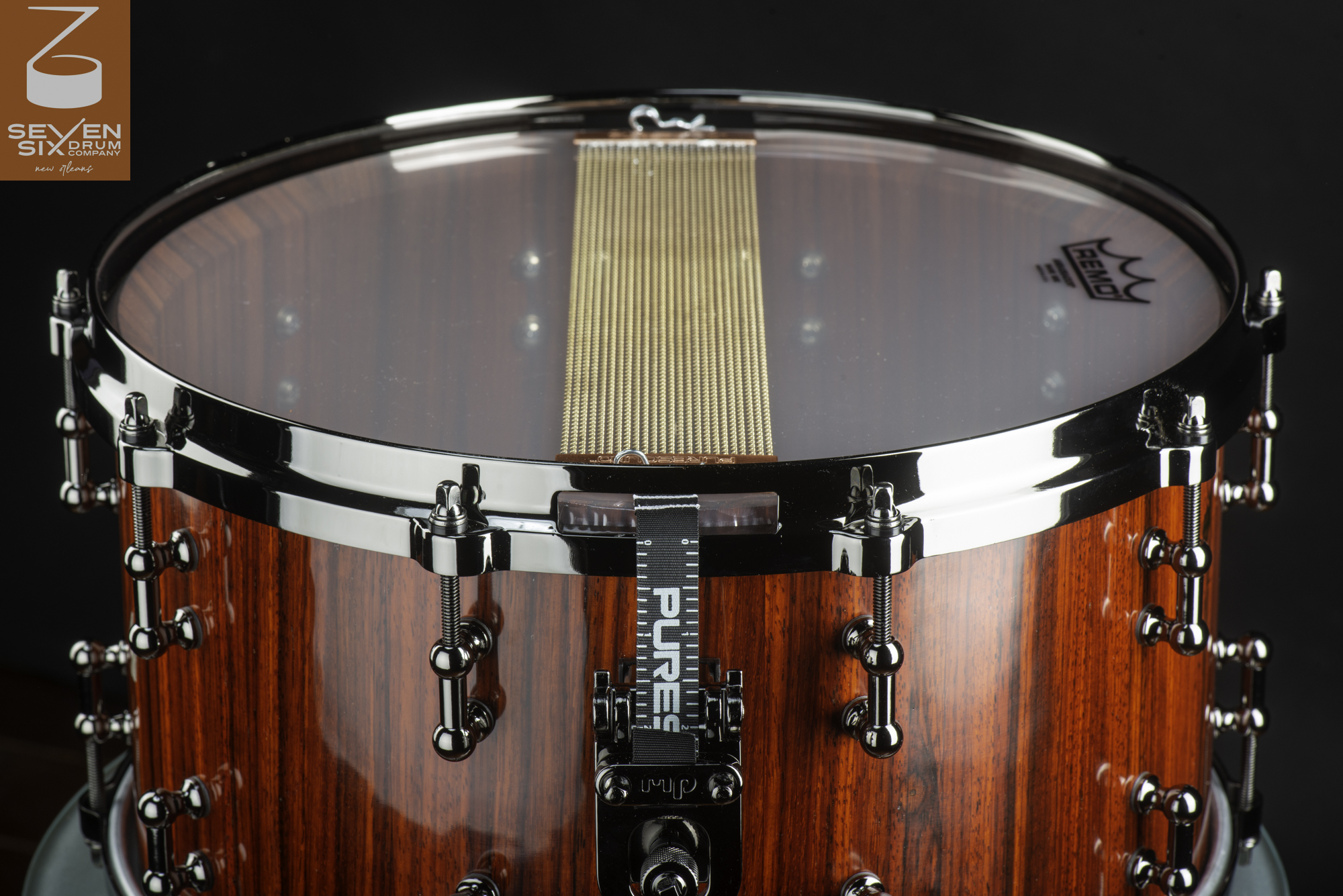 Handmade snare drum featuring a glossy red-brown wood shell, black nickel hardware, and brass snare wires.