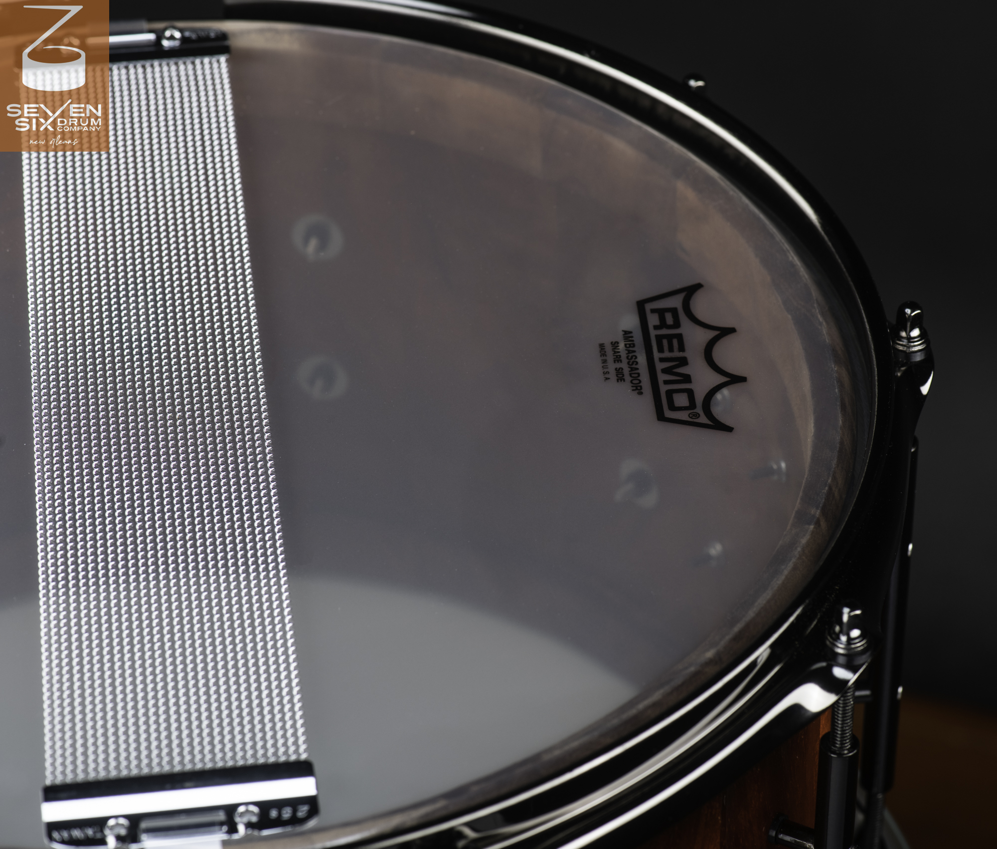 Underside of a reddish-brown wood snare drum with a clear head, silver wire snares, and gloss black metal hoops and lugs.