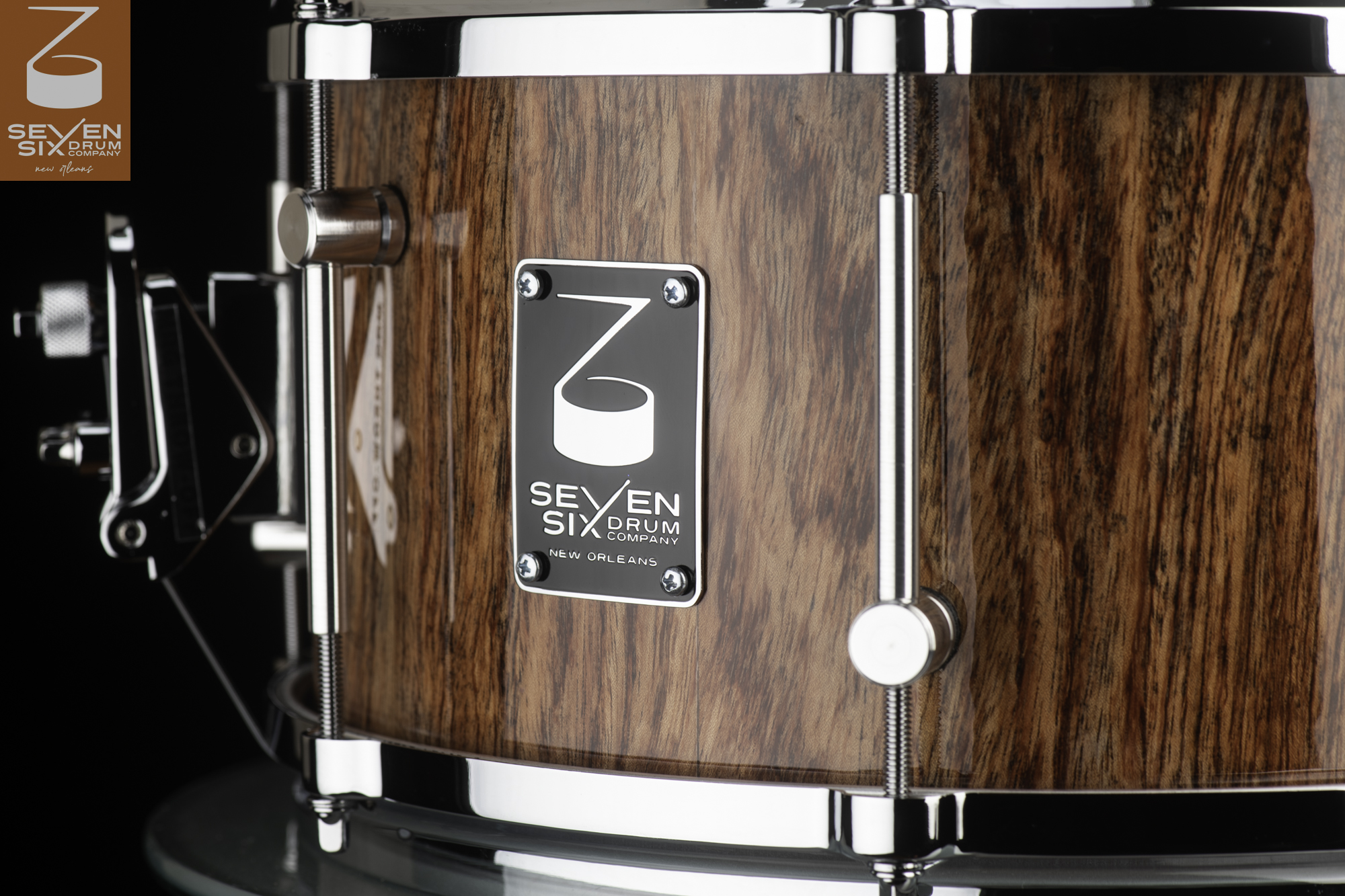 Handmade snare drum with a glossy natural wood shell, polished chrome rims, offset tube lugs, and a black metal logo badge.