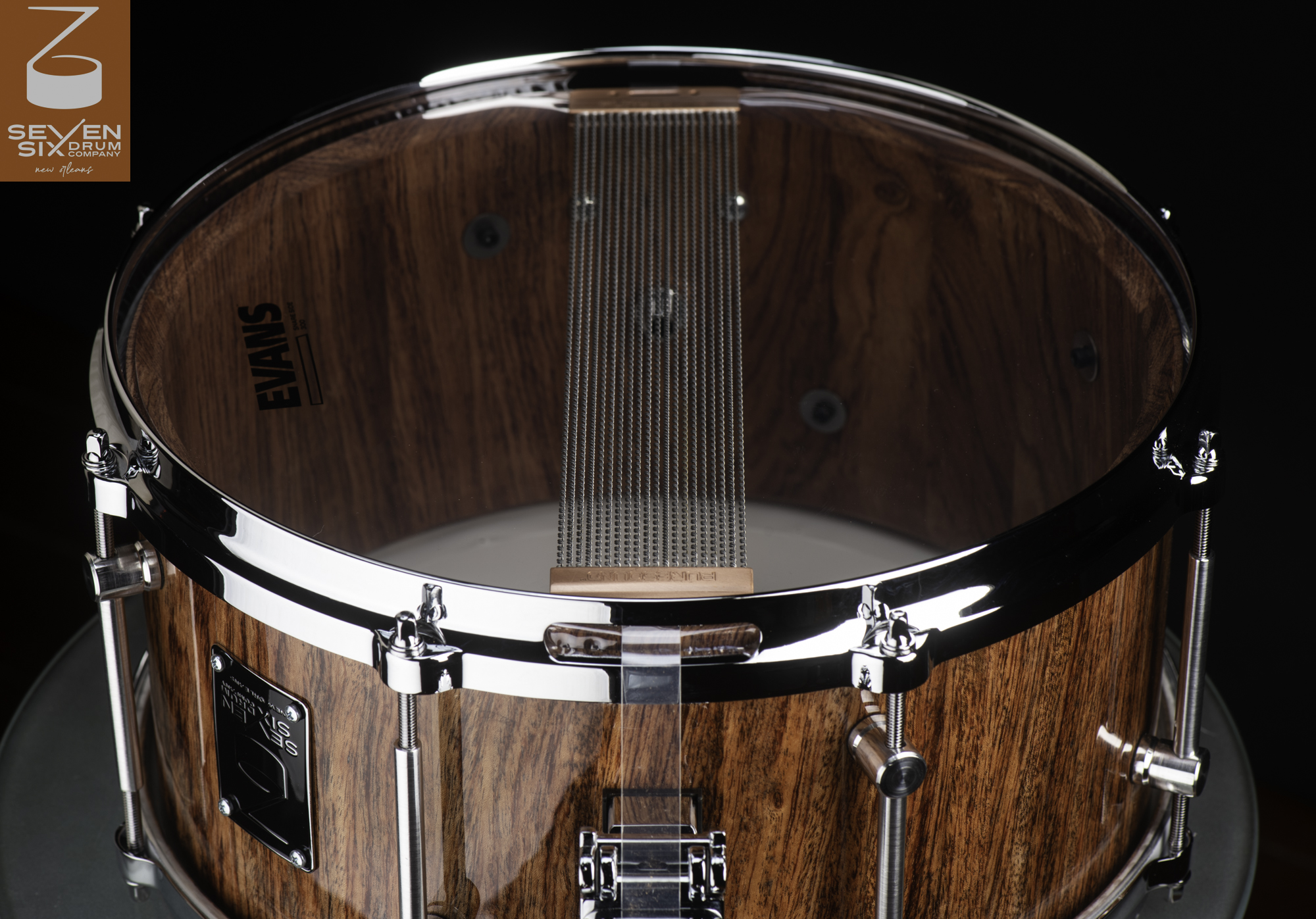 Handmade snare drum with a glossy wood grain shell, chrome hardware, and snare wires across a clear resonant head.