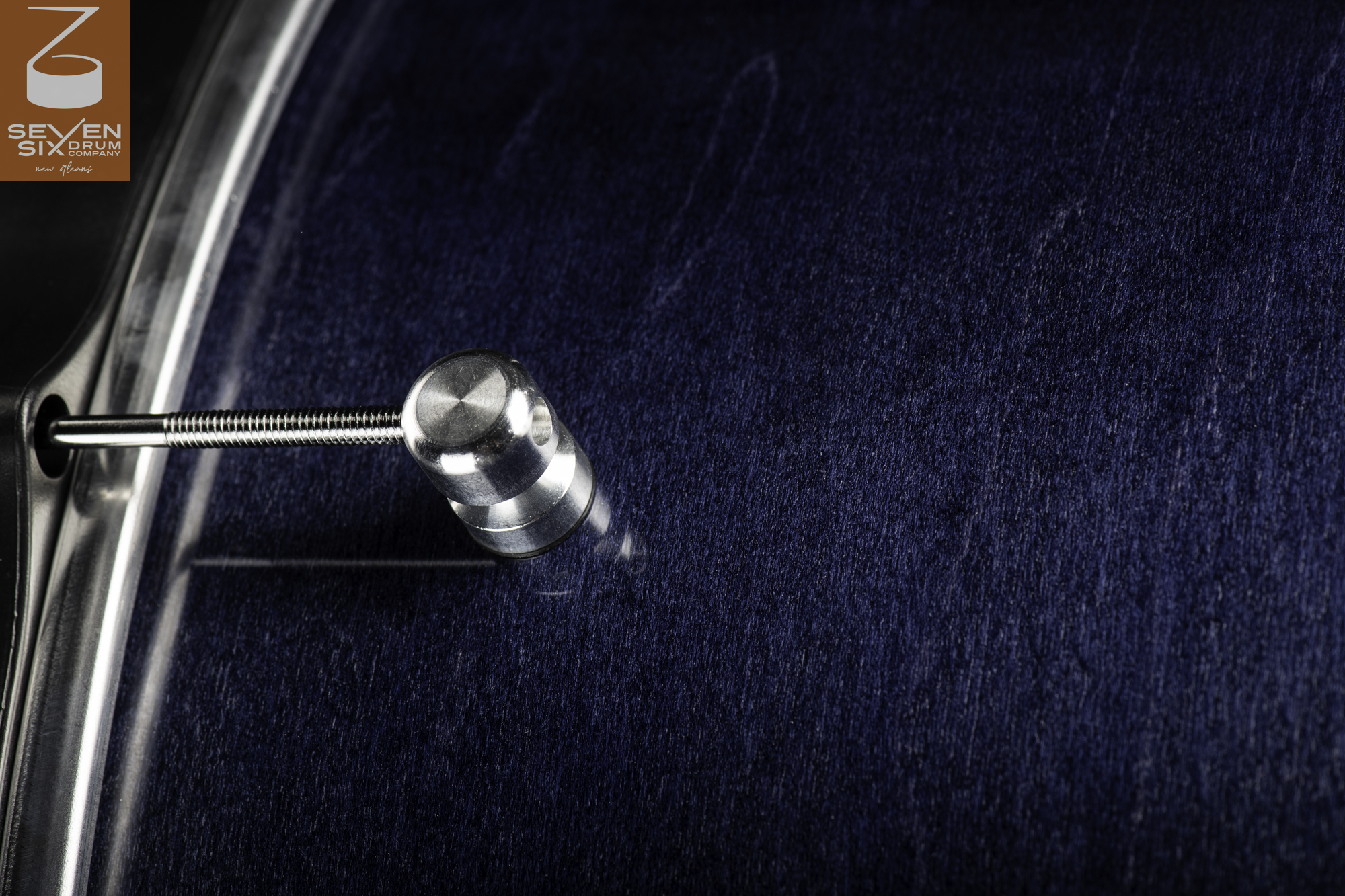 Close-up of a snare drum featuring a dark blue stained wood shell, brushed metal lug, tension rod, and chrome hoop.