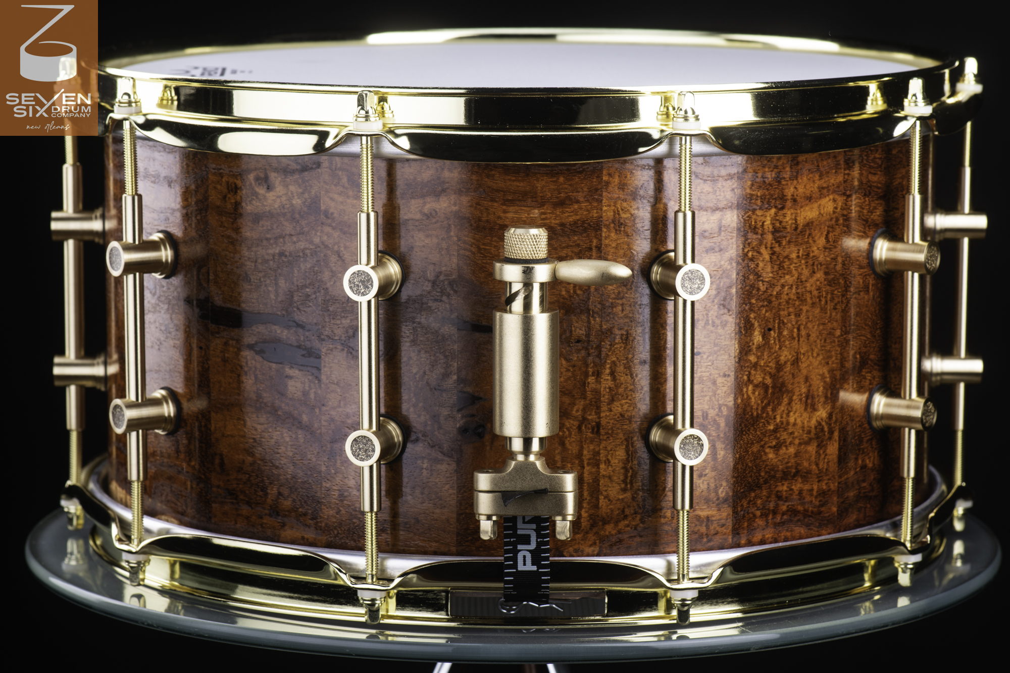 Handmade snare drum with a glossy figured brown wood shell, polished brass hoops, tube lugs, and a brass throw-off.