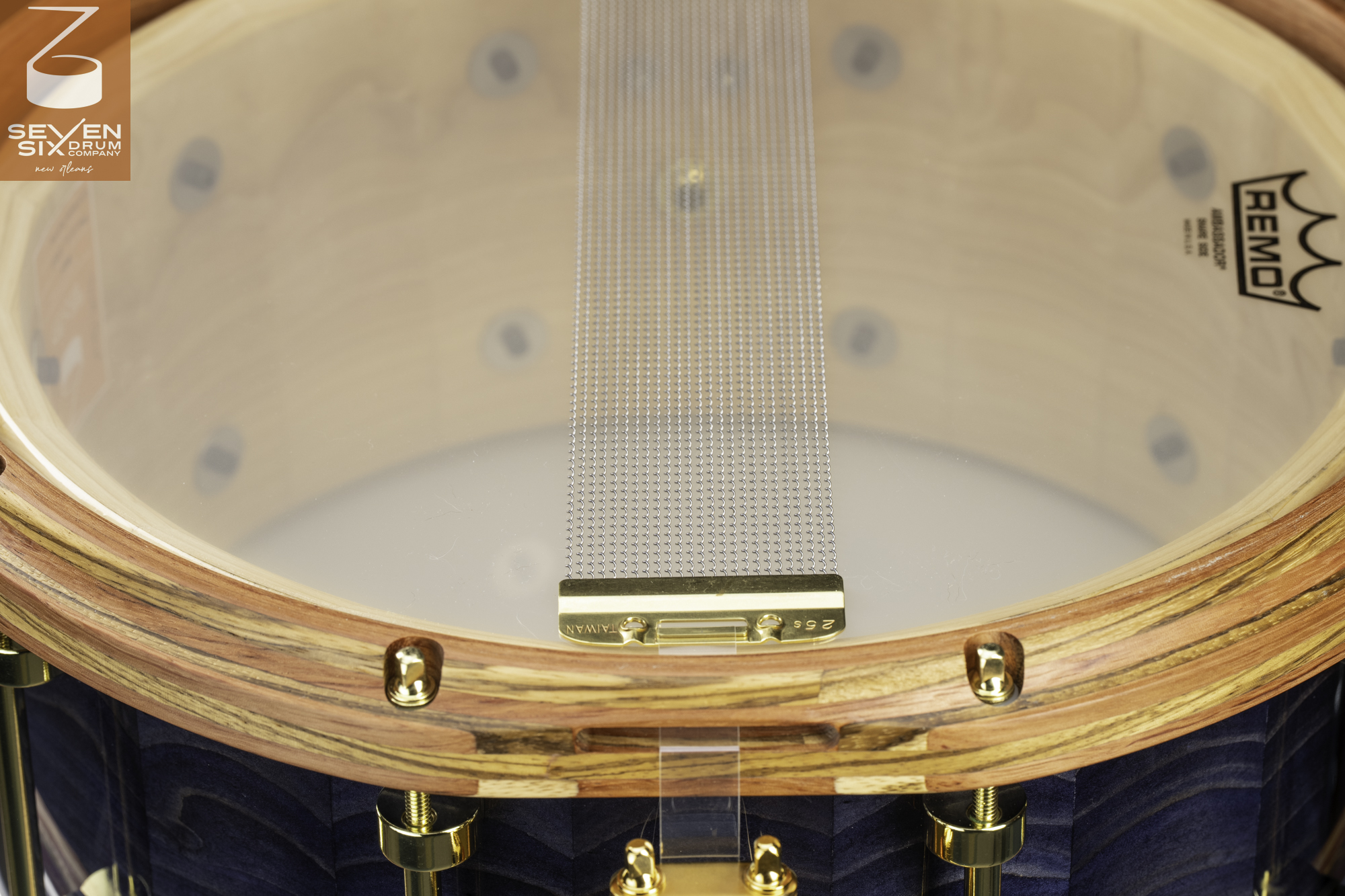 Bottom of a snare drum with wire snares, a clear head, striped wood hoop, blue wooden shell, and polished gold hardware.