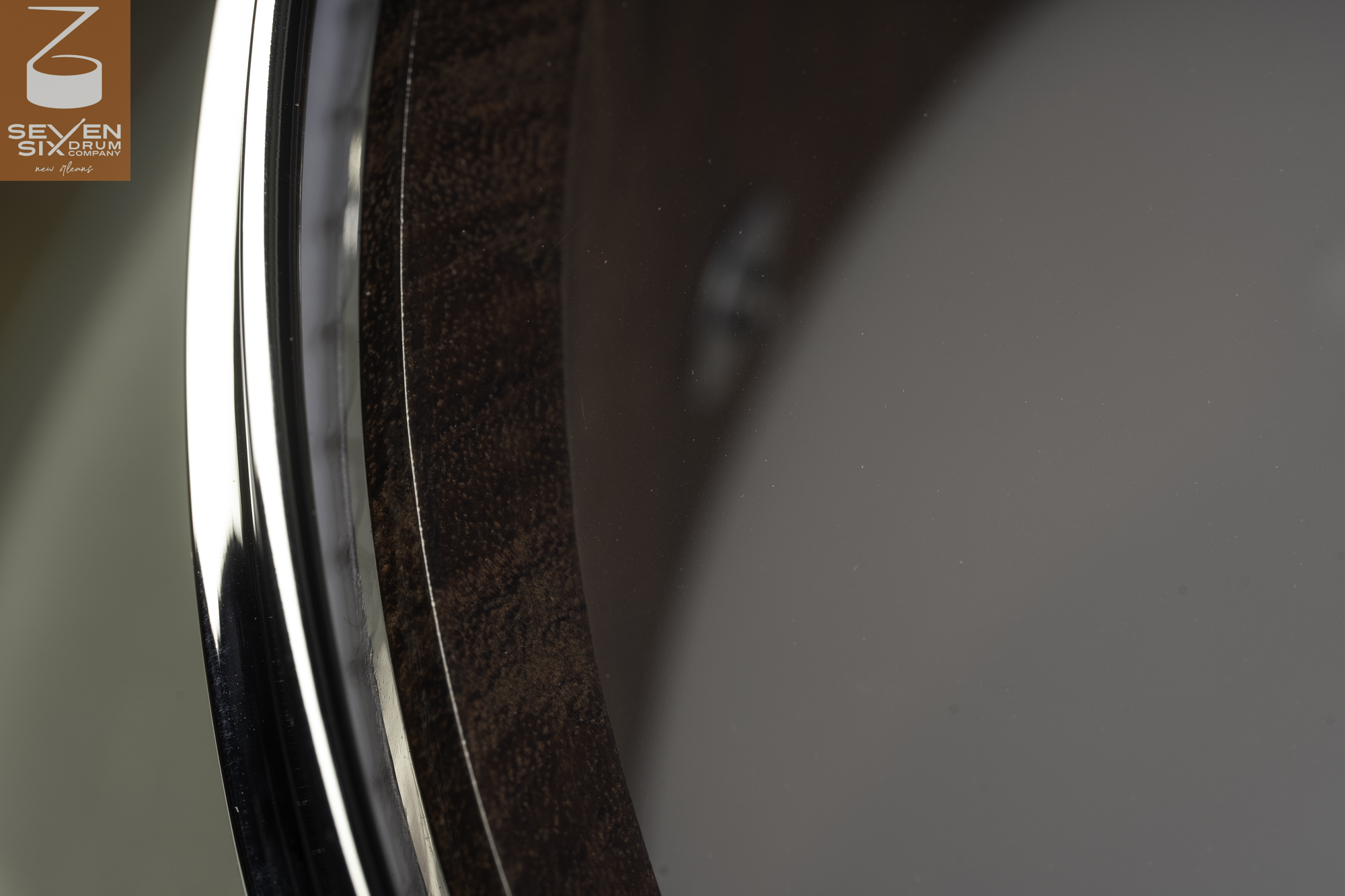 Detail of a dark stained wood snare drum shell with a smooth finish, clear head, and polished chrome hoop.