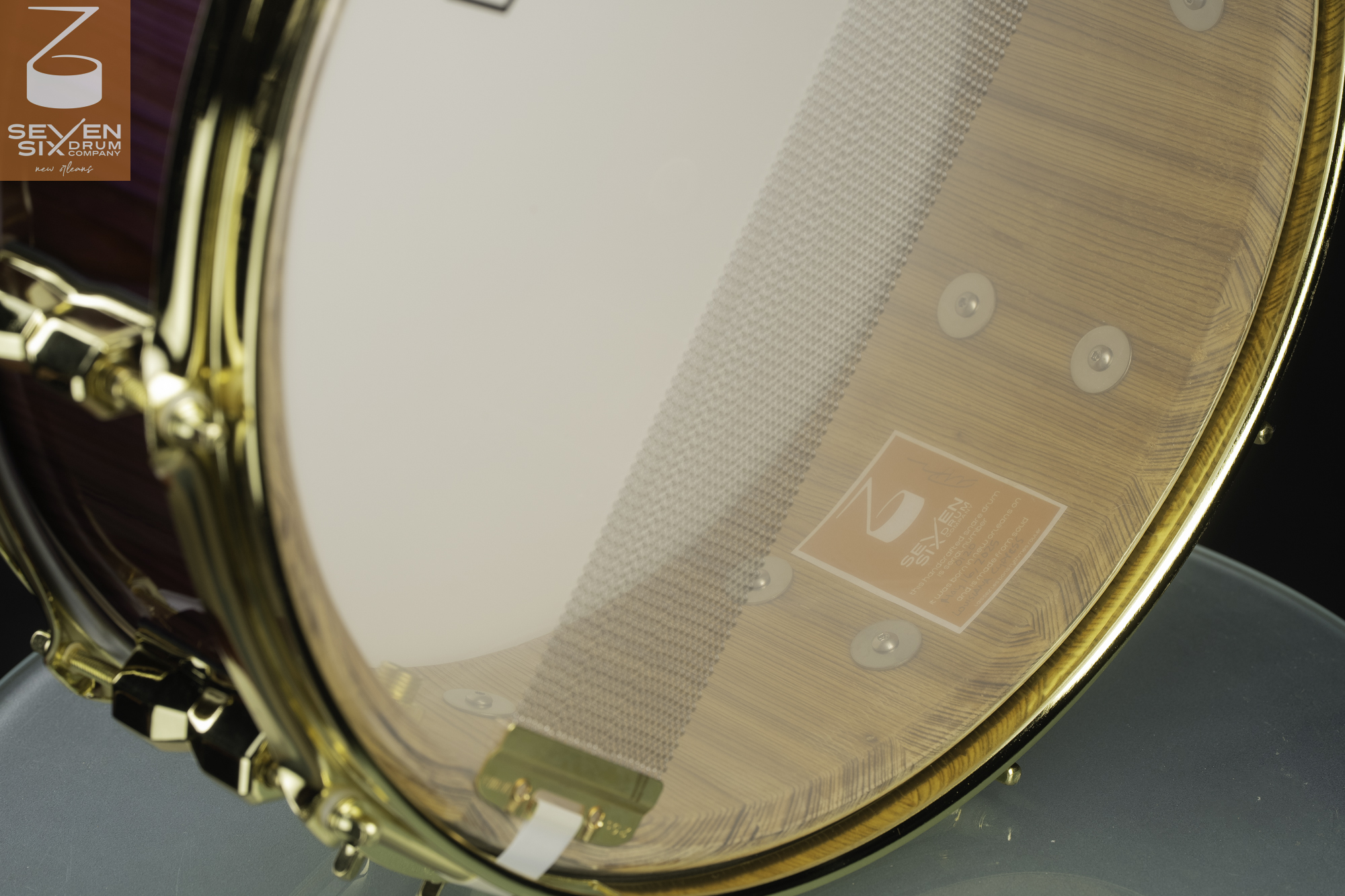 Underside of a wooden snare drum showing a natural grain interior, snare wires on a clear head, and polished gold hardware.