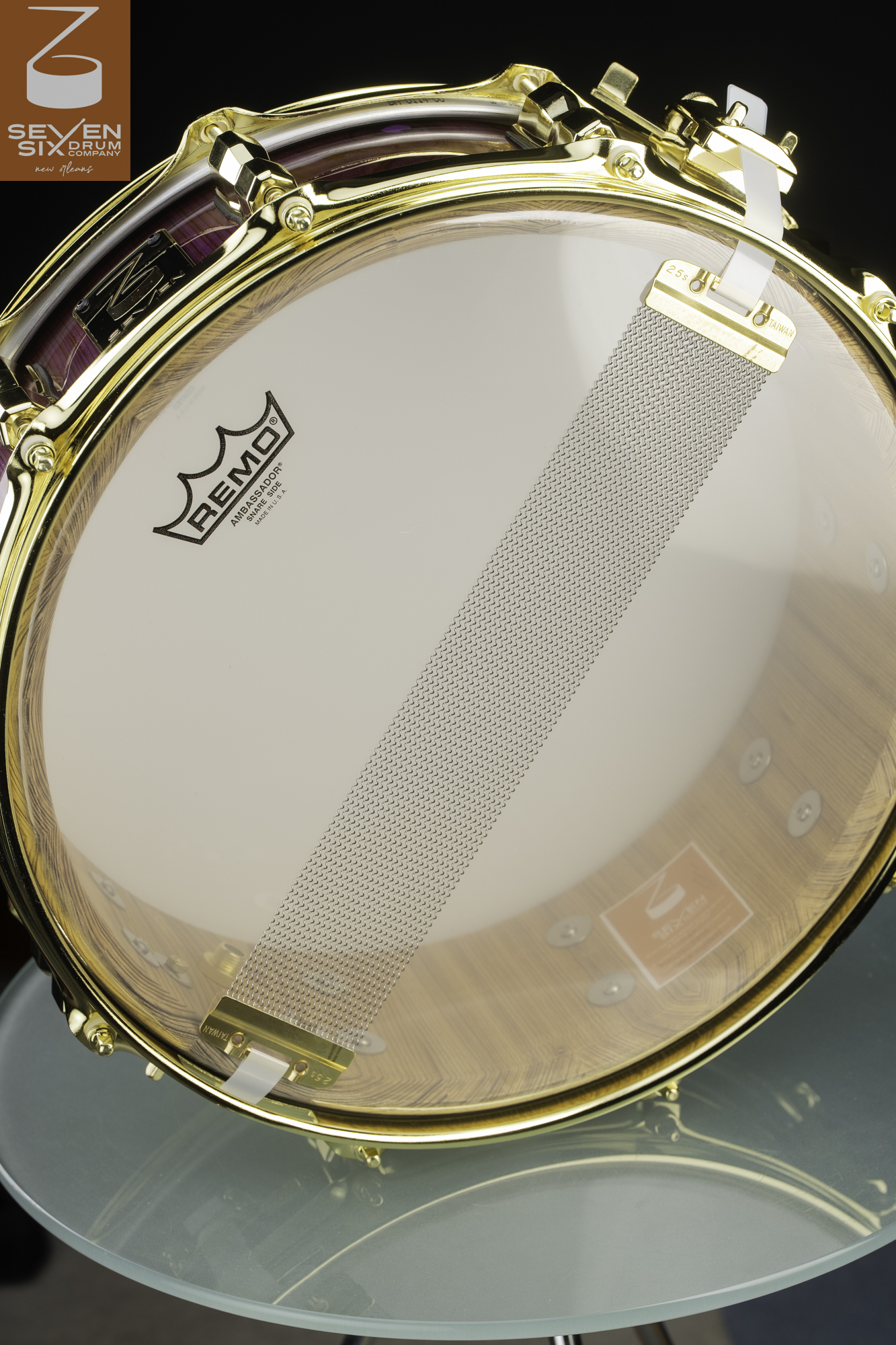 Snare side of a drum showing a natural wood grain interior, purple exterior finish, gold hardware, and steel snare wires.
